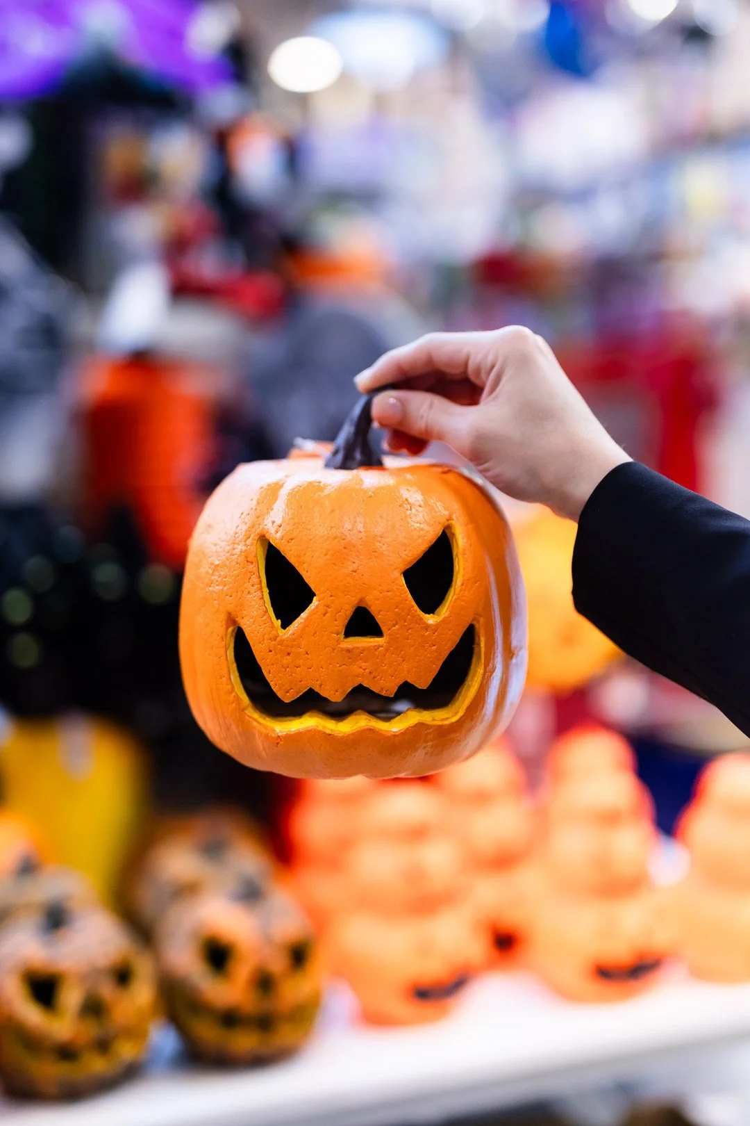 Person holding a pumpkin-shaped plastic lantern with a jack-o'-lantern face displayed at a Halloween or fall market.