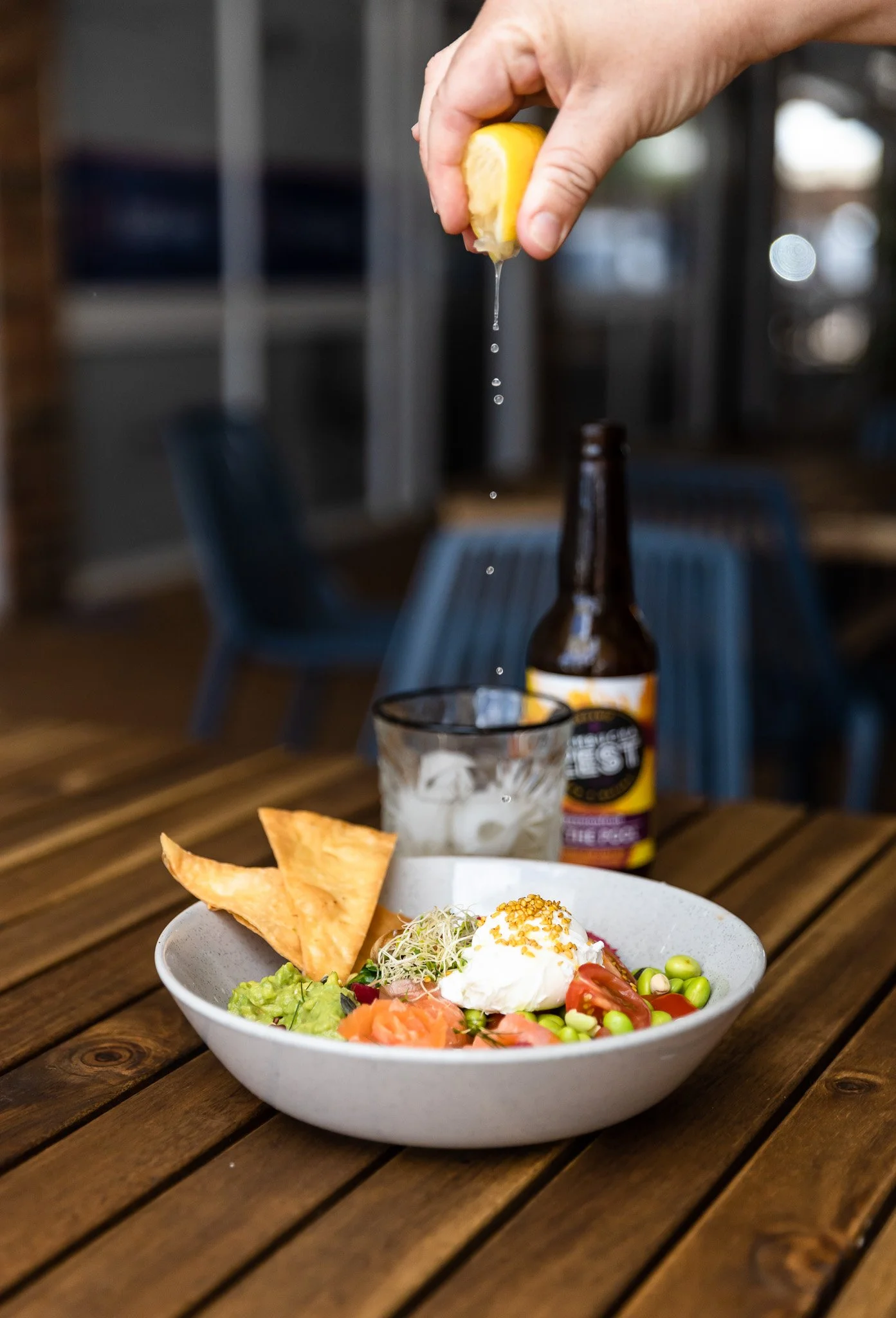 A hand squeezing a lemon over a salad in a bowl on a wooden table, with beer, ice, and a glass in the background.