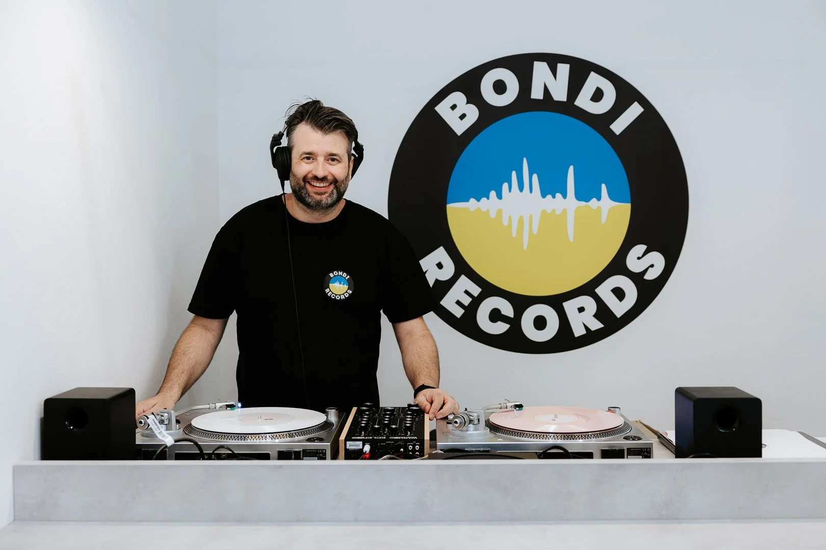 Craig Fordham, the owner of Bondi Records, standing behind the vinyl decks in the shop with a large Bondi Records logo on the wall behind him