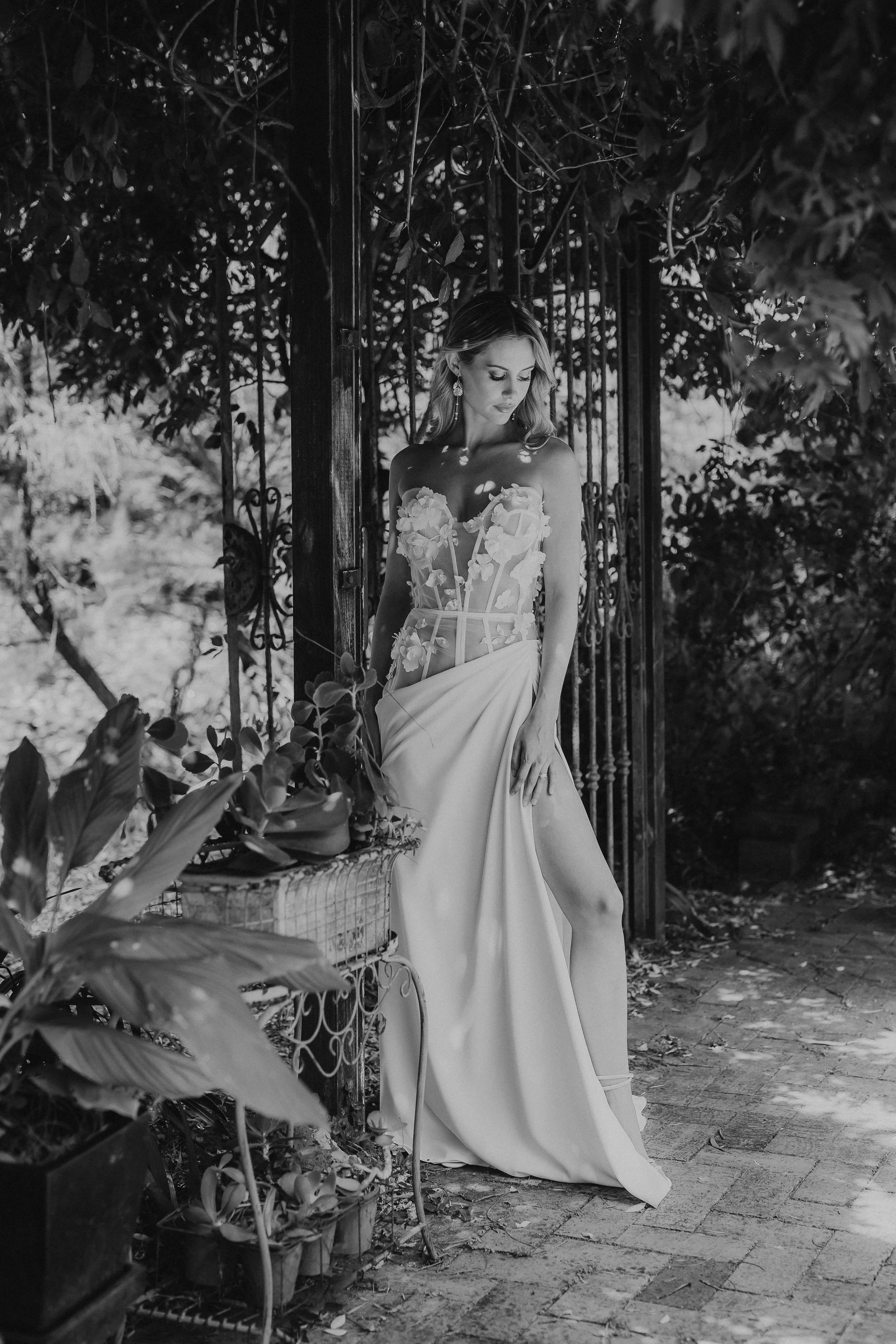 A woman in a floral wedding dress stands outdoors near a wrought iron gate and lush foliage, posing gracefully with her eyes downcast.