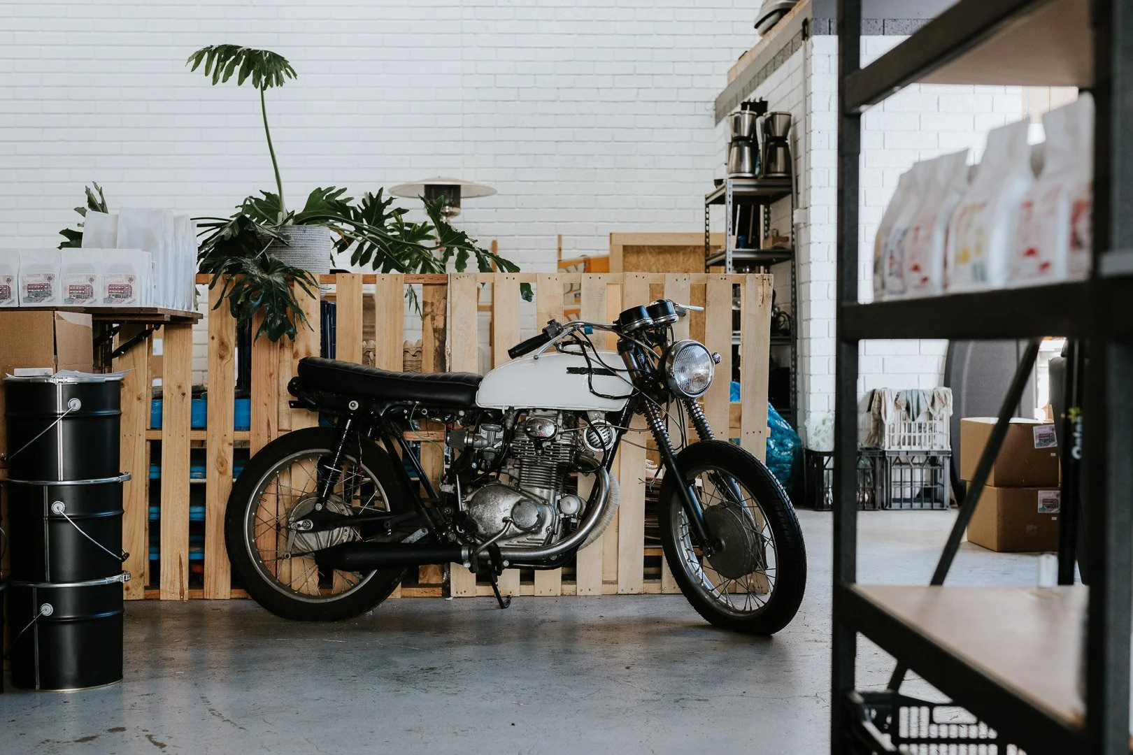 A vintage motorcycle parked inside a store or warehouse with white brick walls and wooden pallets, surrounded by shelving units and boxes.
