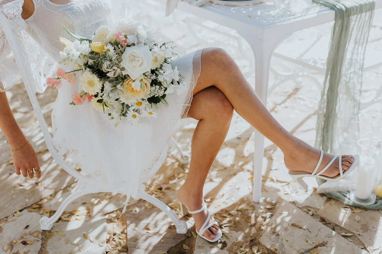 Part of a woman sitting on a white chair, holding a bouquet of white, yellow, and pink flowers, wearing a white lace dress, and white high-heeled sandals, with a white table nearby on a sandy area covered with fallen leaves.