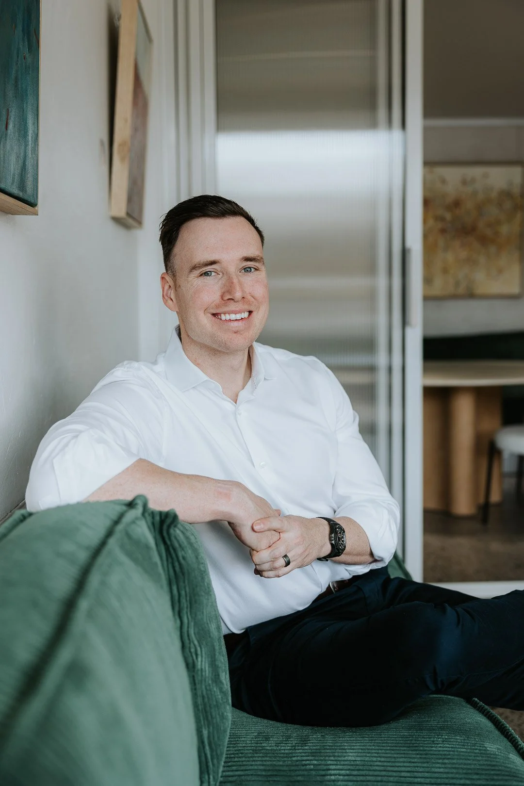 Relaxed Corporate Headshots and Team photography on the Central Coast. A man in a business shirt, sitting on a bench seat in an office in Terrigal, looking sideways towards the camera, looking comfortable and smiling.