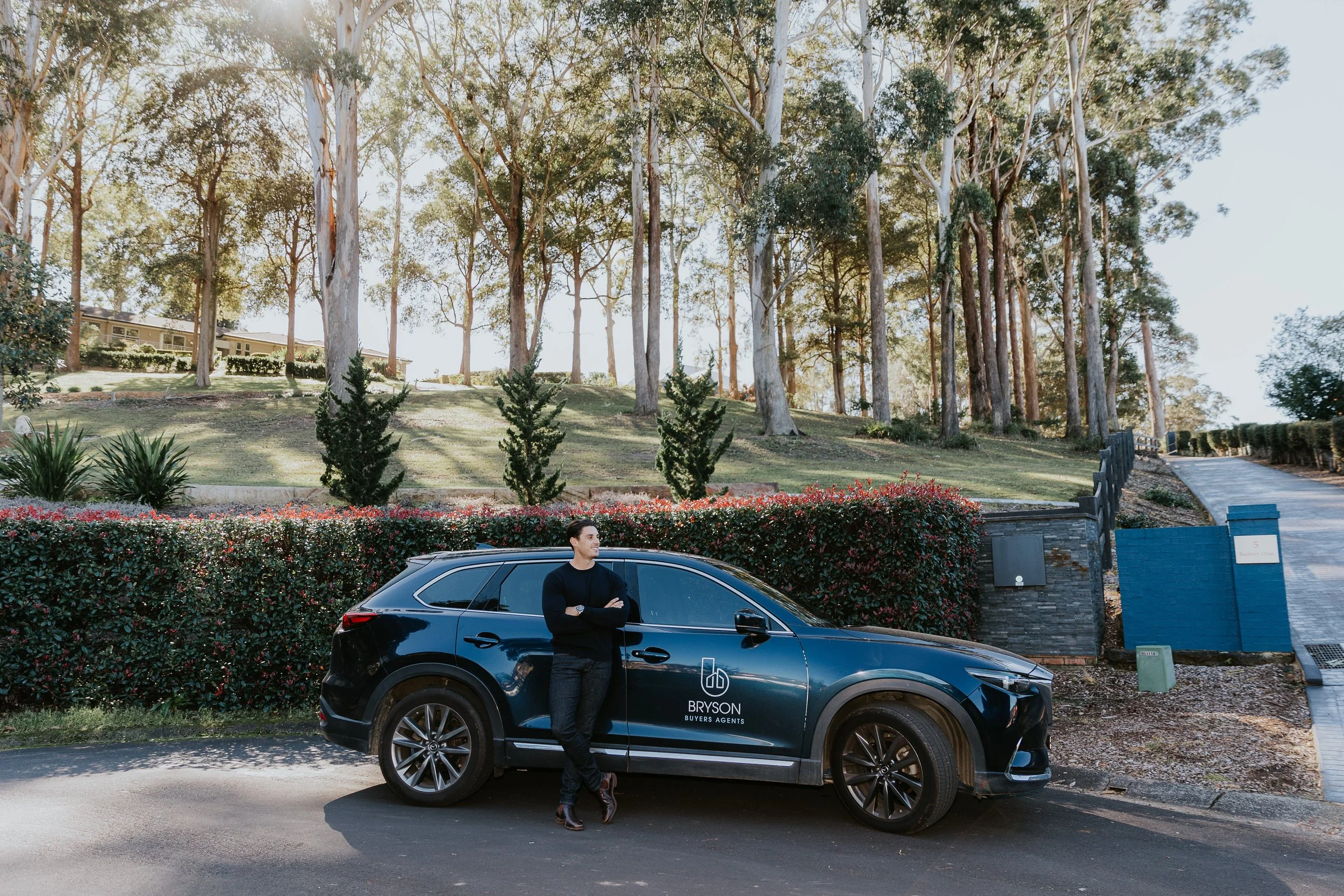 A man standing next to a black SUV with 'Bryson Buyers Agents' written on the side, parked on a street with a hilly landscape and tall trees in the background.