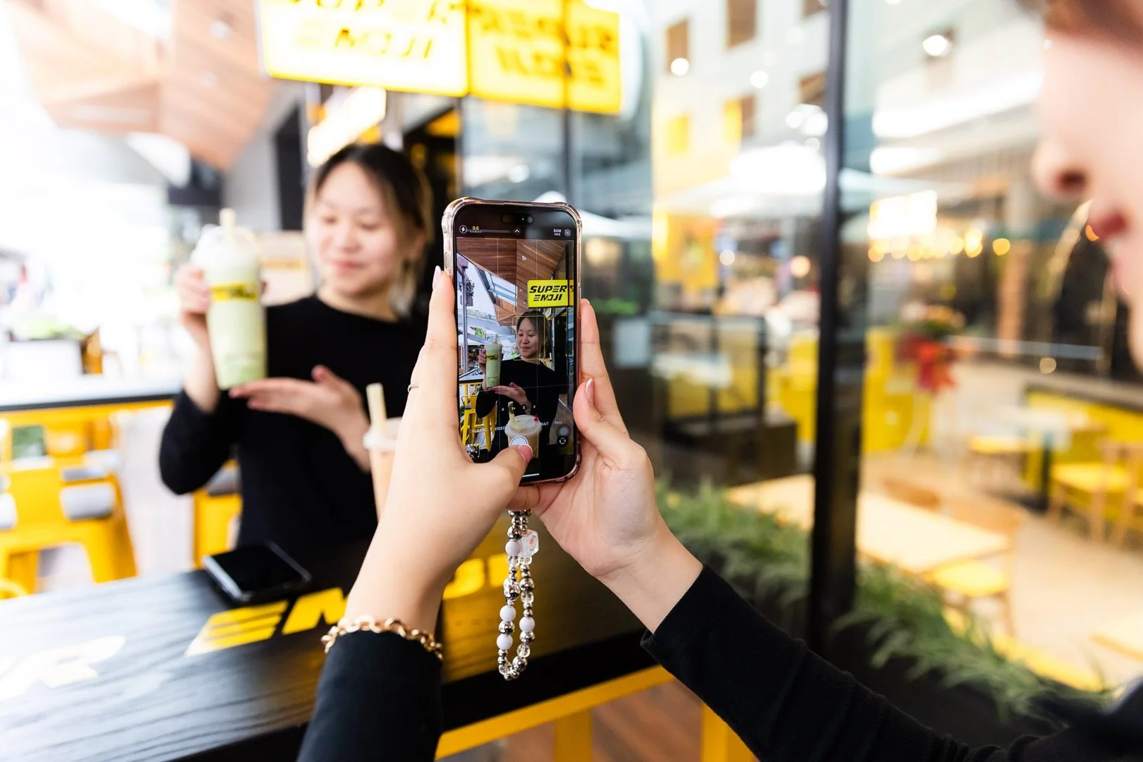 Person taking a photo of a woman holding a drink outside a cafe with yellow decor