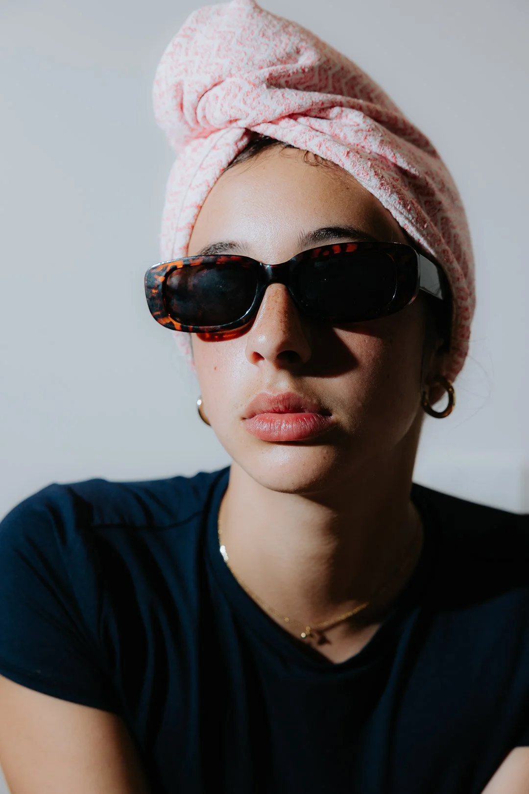 A woman with fair skin wearing dark sunglasses, a pink towel wrapped around her head, gold hoop earrings, a small gold necklace, and a black shirt, against a plain light background.