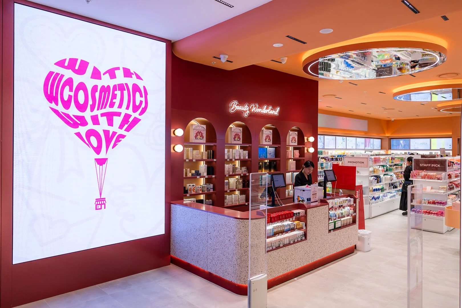 Inside a colorful retail store with a red and beige color scheme, a large digital screen displays a pink heart-shaped balloon with the words 'Love with WOSMETIC' written on it. The store has a checkout counter with two staff members, shelves stocked 