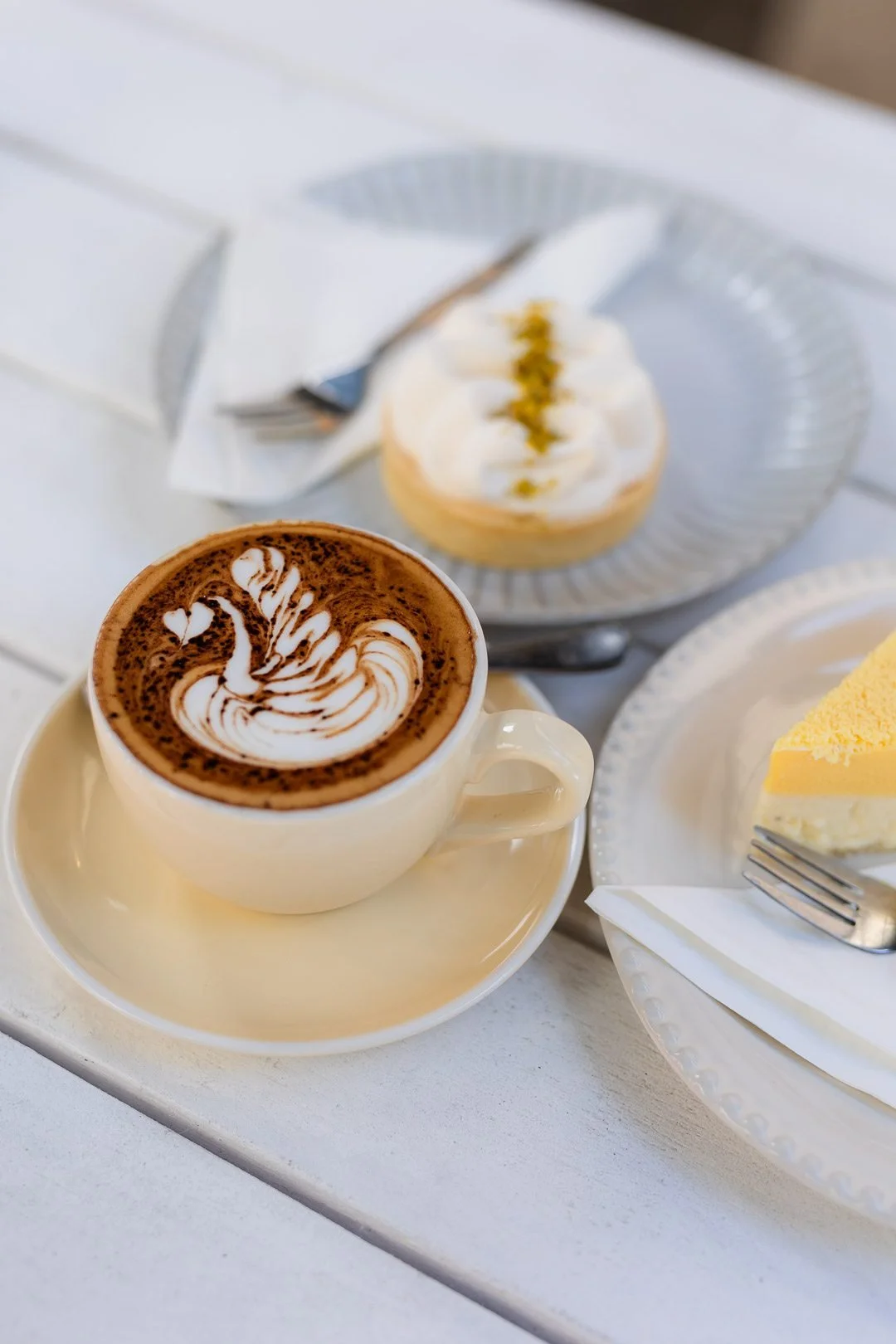 A cup of hot chocolate with swirled cream art, a slice of lemon cheesecake, and a lemon meringue pie on white plates and a table.