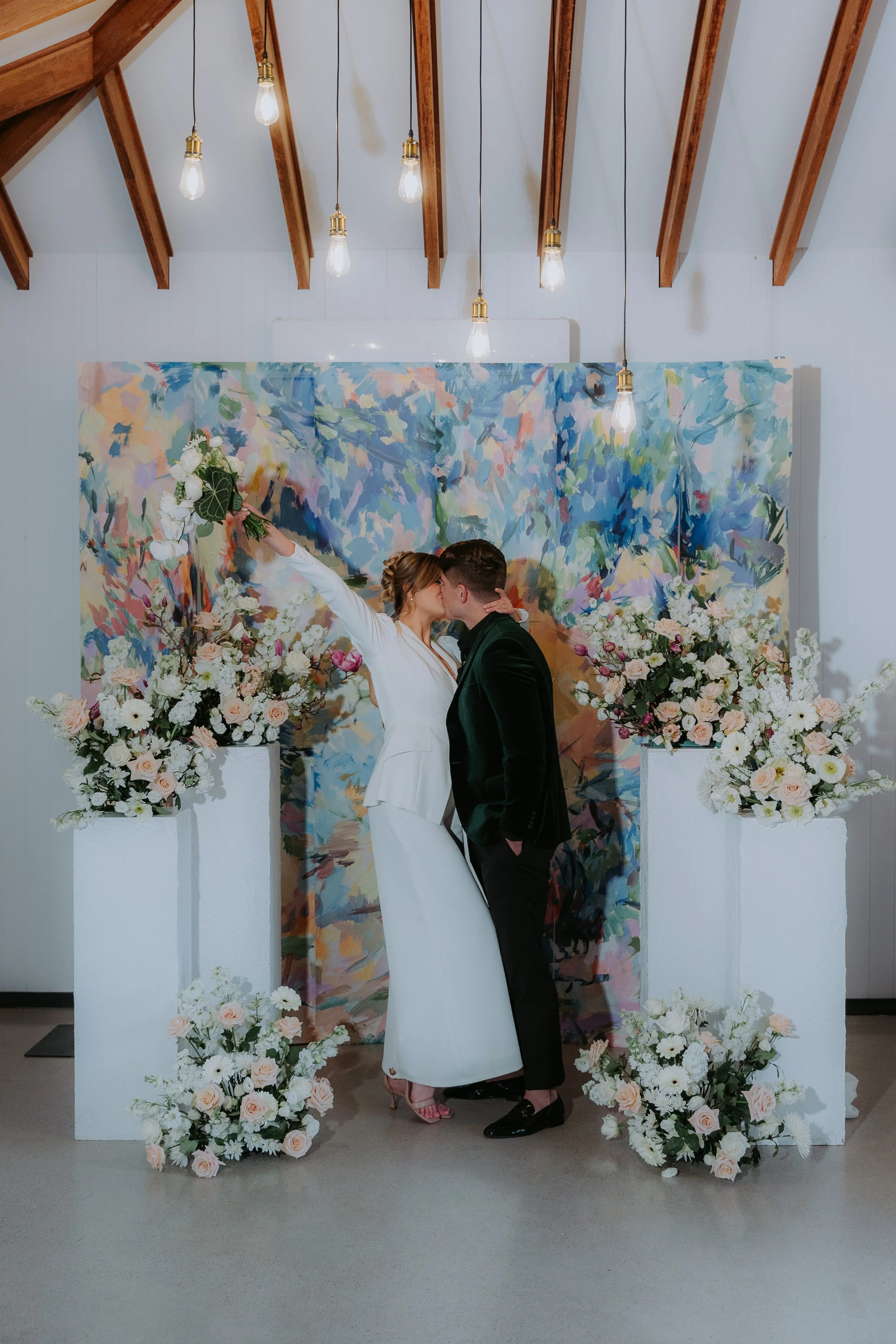 A couple kissing at their wedding ceremony in front of a colorful abstract backdrop with floral arrangements.