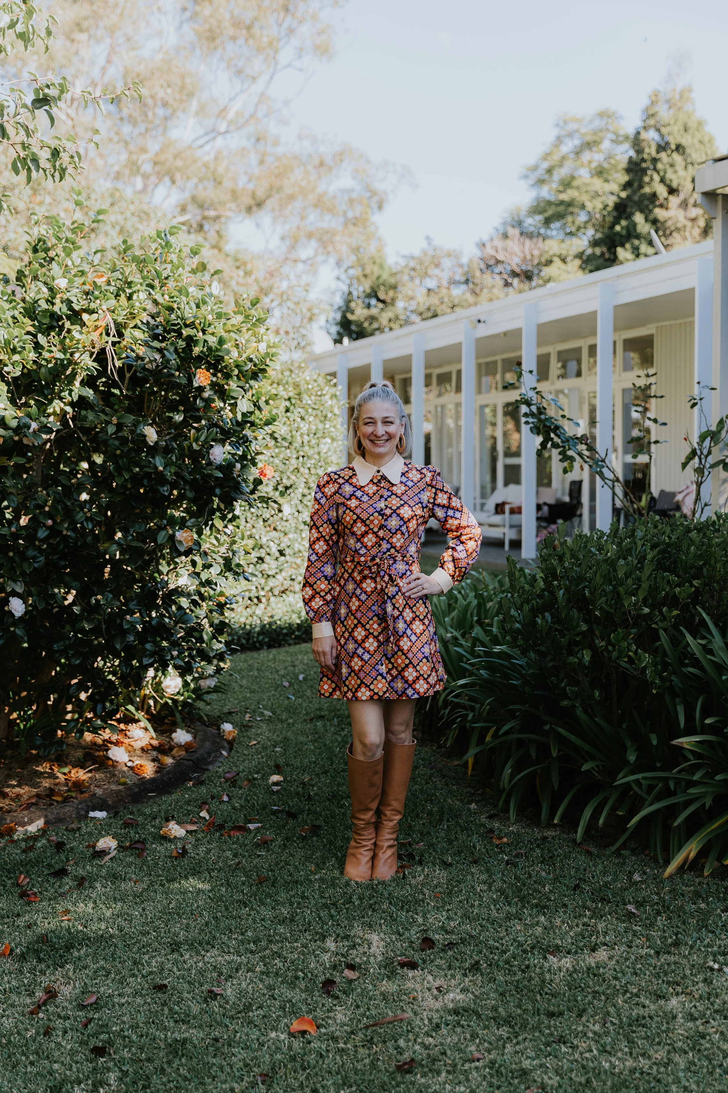 A woman standing on a grassy yard in front of bushes and a house, smiling and wearing a colorful patterned dress and tall brown boots.