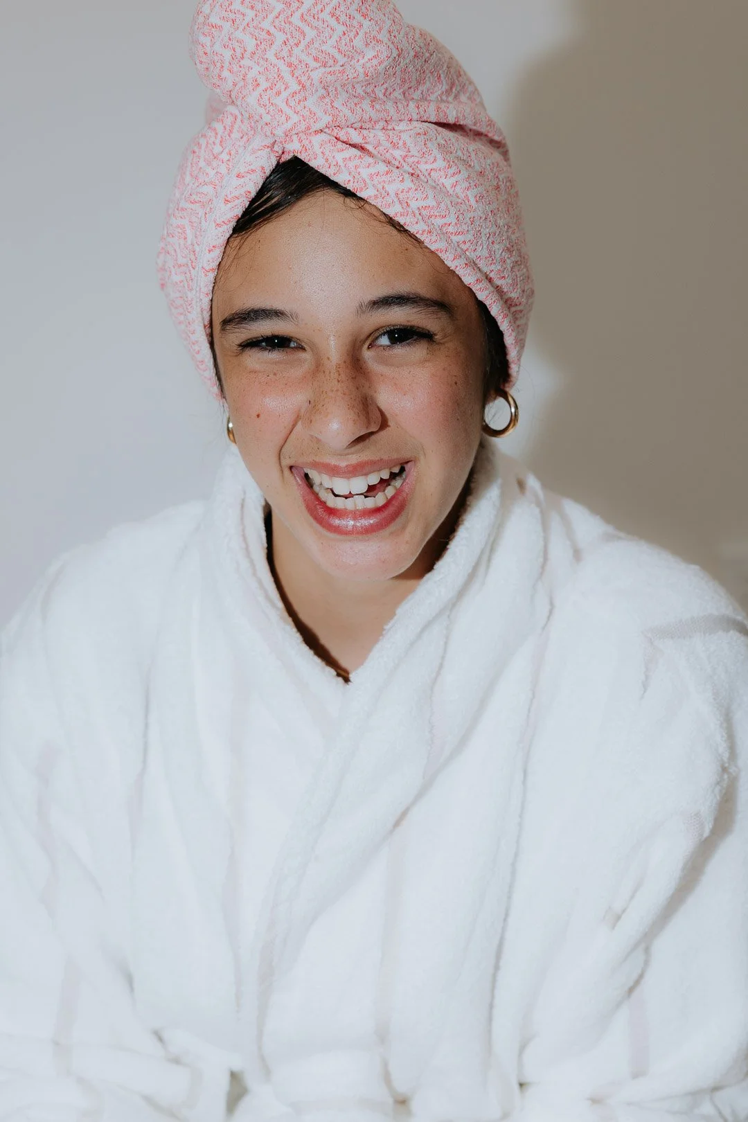Woman with a pink towel wrapped around her head, smiling and wearing a white bathrobe.