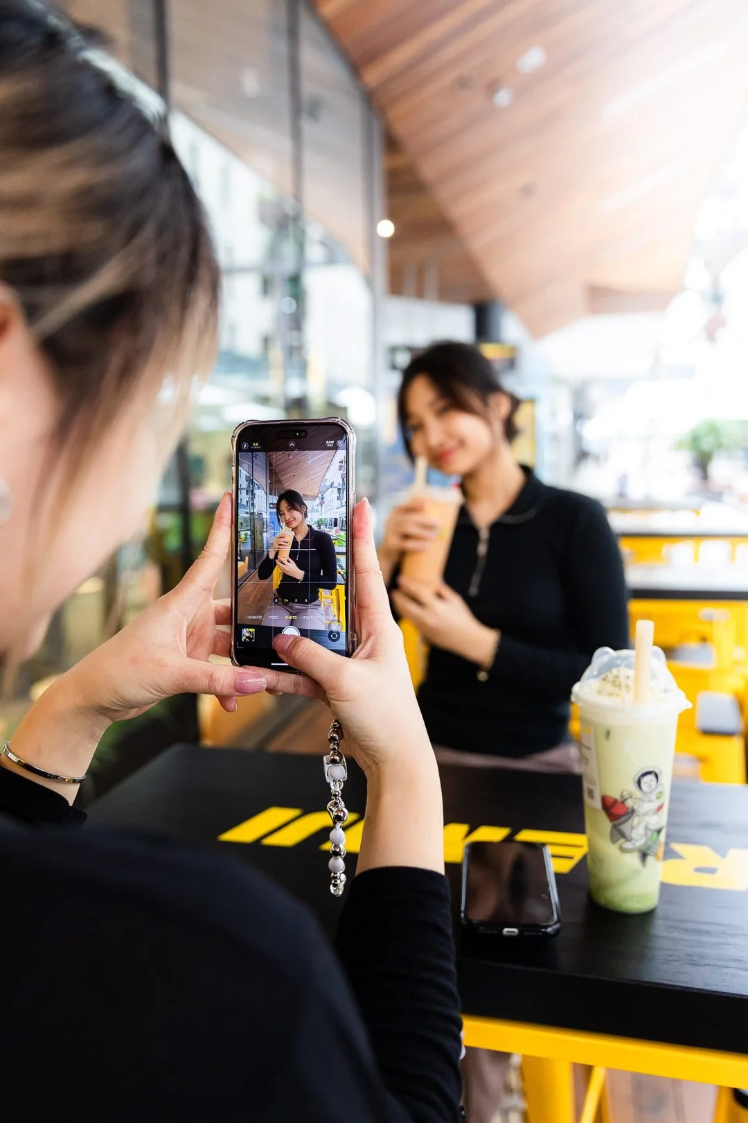 A woman is taking a photo of another woman in a cafe with a phone. The woman in the background is smiling and holding a drink, with a beverage on the table nearby.