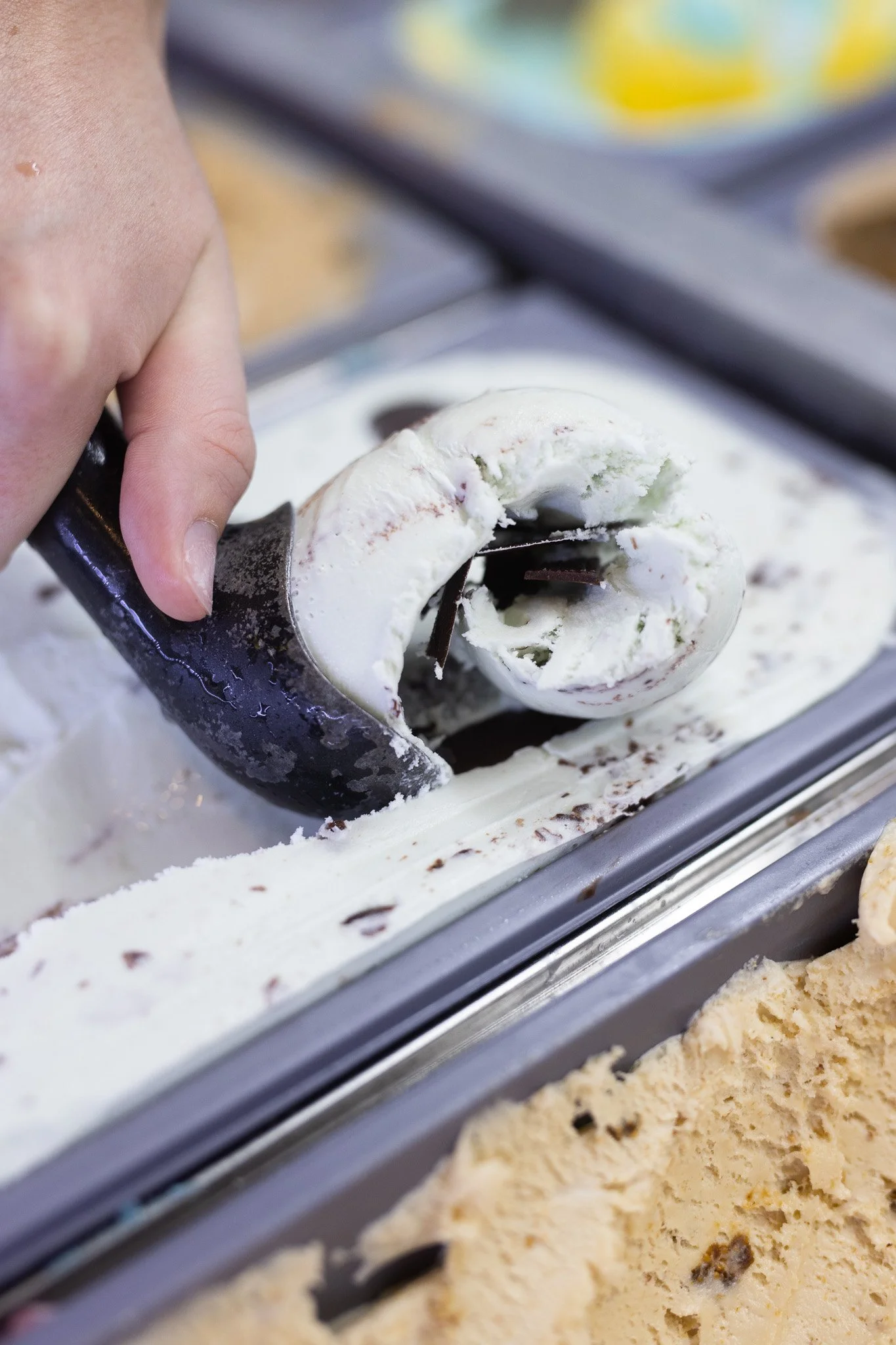 Person scooping vanilla ice cream with chocolate chips from a metal container at an ice cream shop.