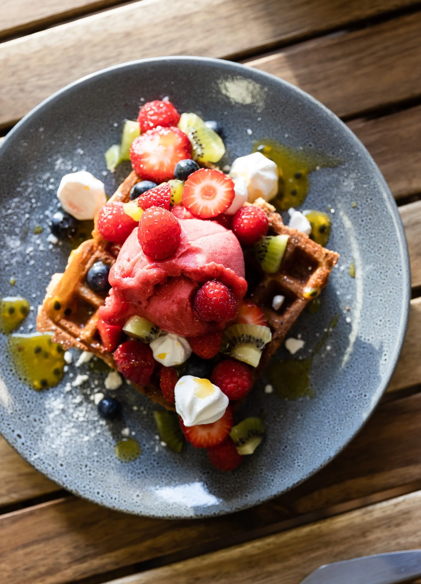 Waffle topped with mixed berries, a scoop of pink sorbet, whipped cream, and drizzled with syrup and honey on a dark gray plate. Wooden table surface underneath.