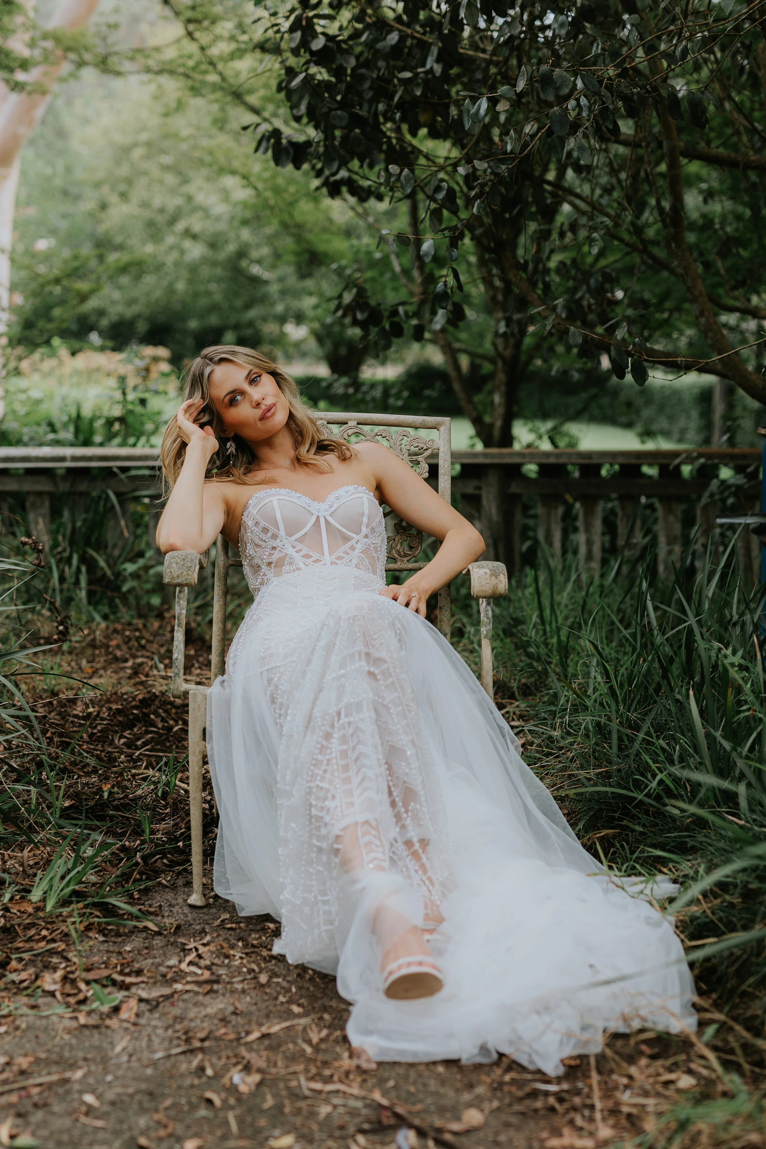 A woman in a white lace dress sits on a garden bench surrounded by greenery, with one hand on her head and a relaxed expression.