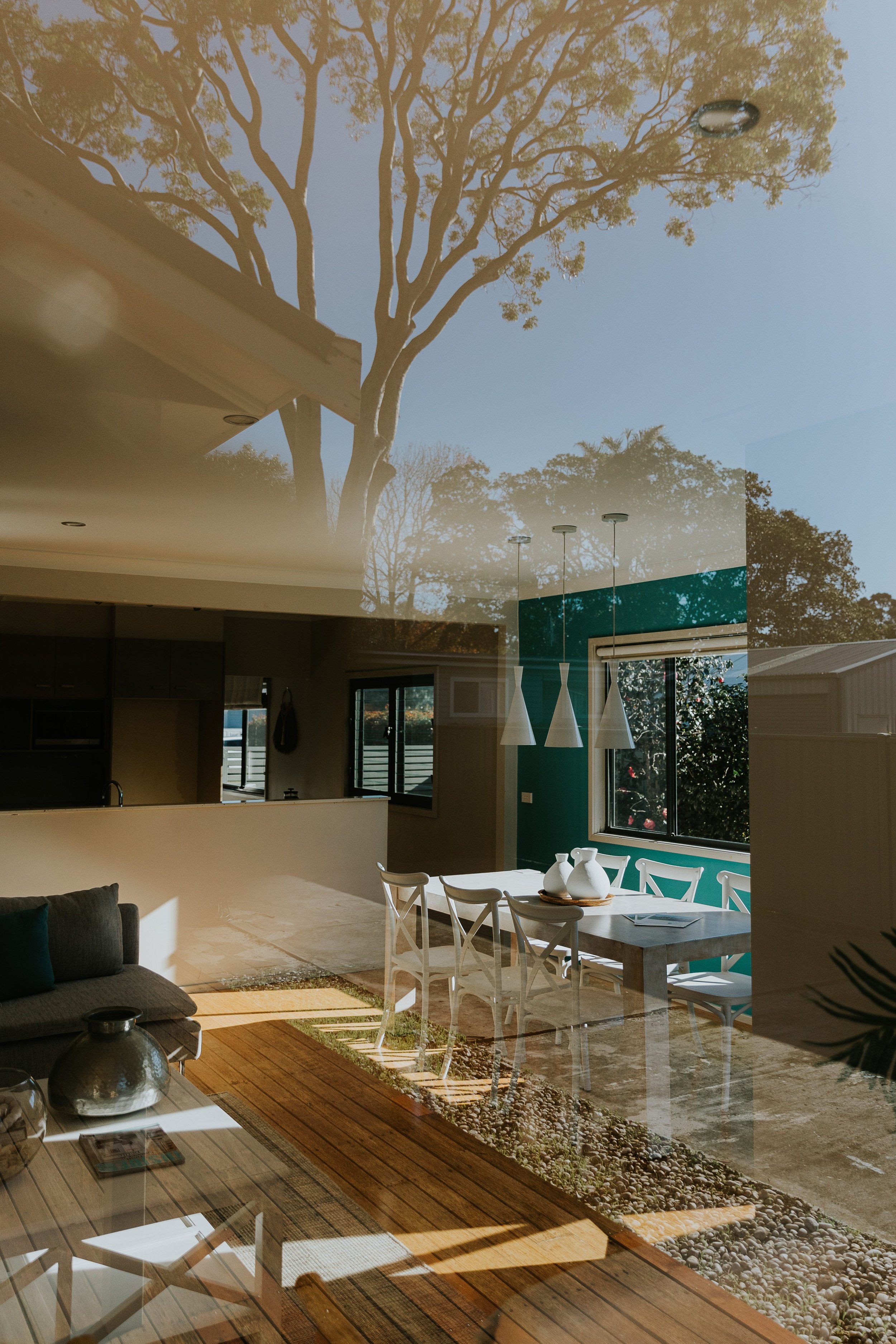 Reflection of dining area with white chairs, white vases on table, teal wall, hanging pendant lights, and large window with outside view, overlaid with the sky and trees from outside.