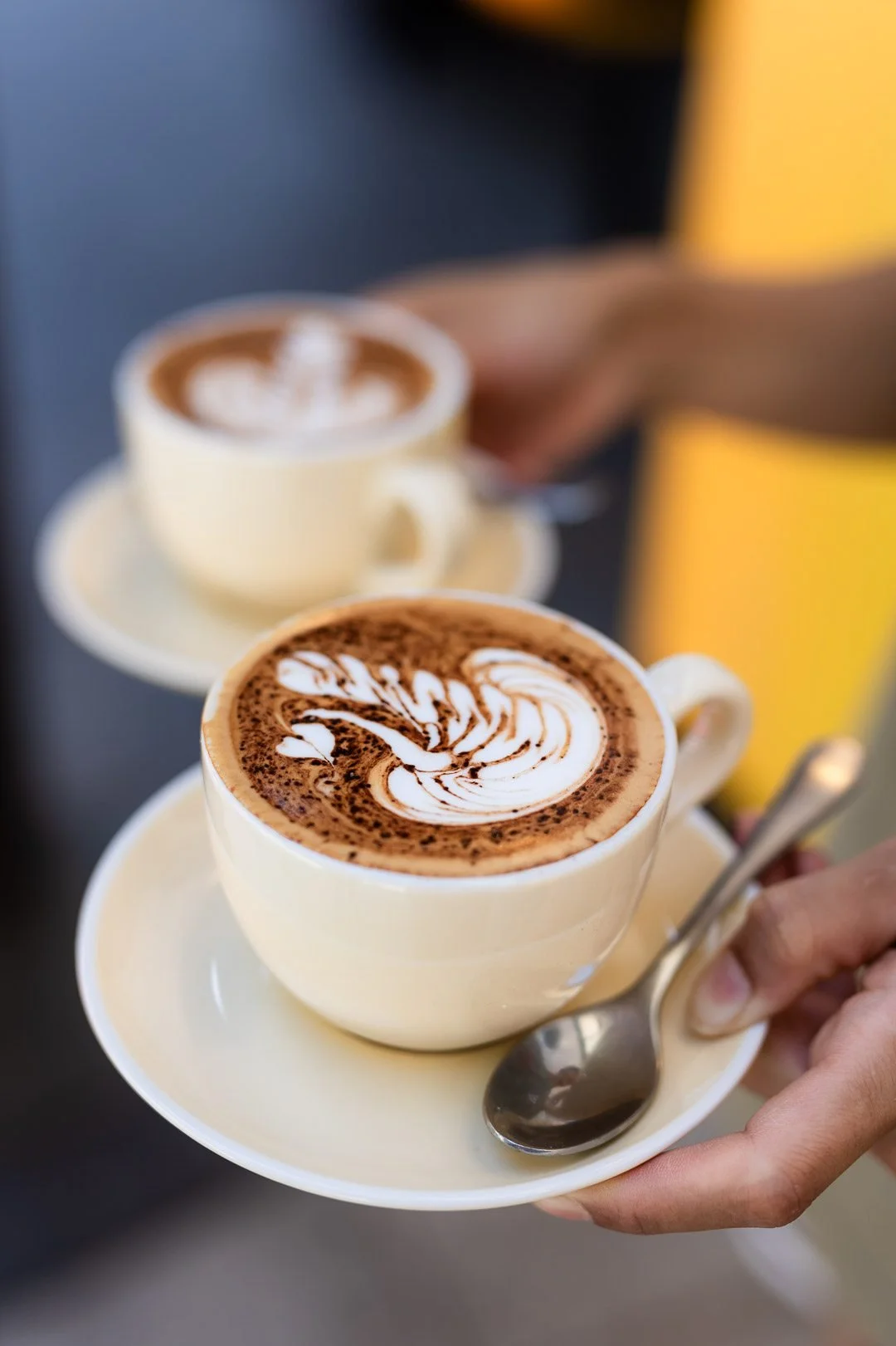 Two cups of cappuccino with latte art, held on saucers with teaspoons, one in foreground and one in background, on a dark surface.