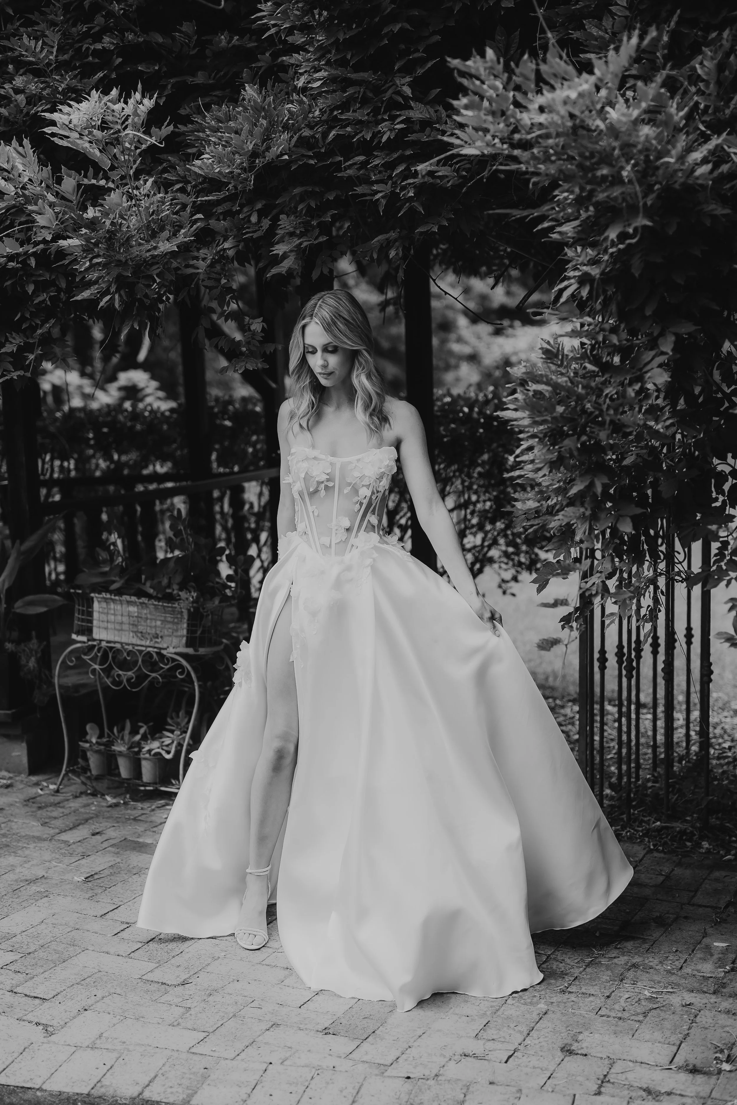 A woman in a wedding dress standing outdoors on a brick path, surrounded by trees and bushes, black and white photograph.