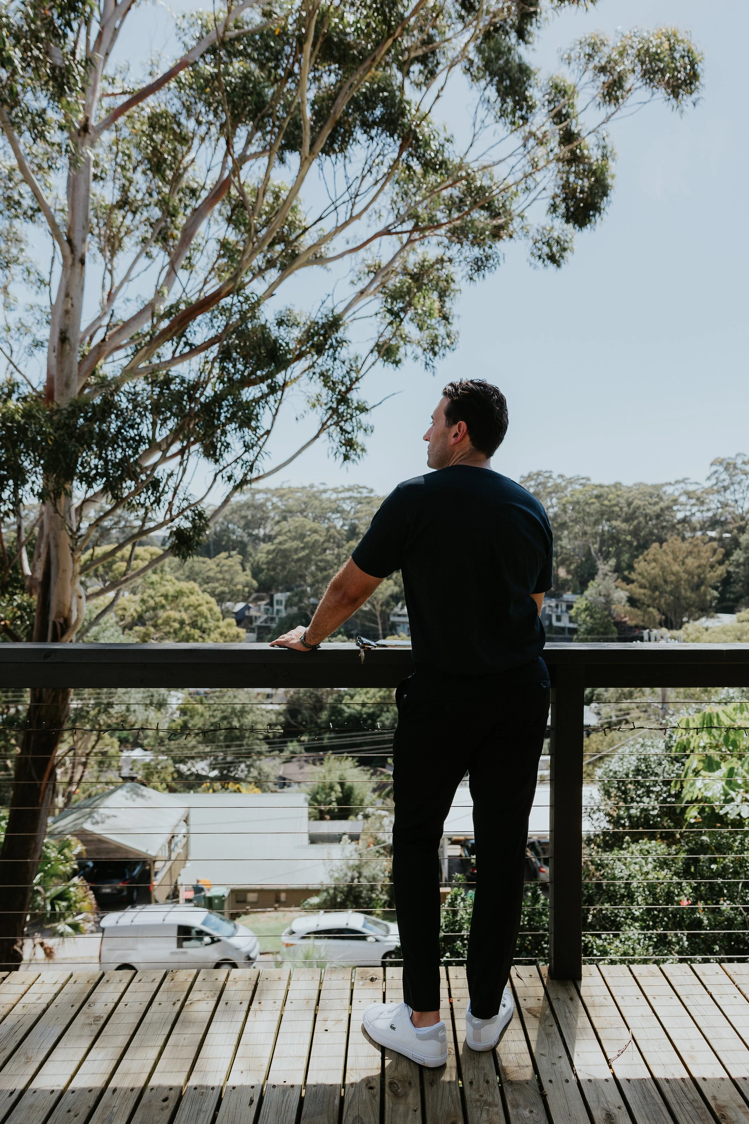 Man in black clothing and white sneakers standing on a wooden balcony, looking out over a neighborhood with trees and houses under a clear blue sky.