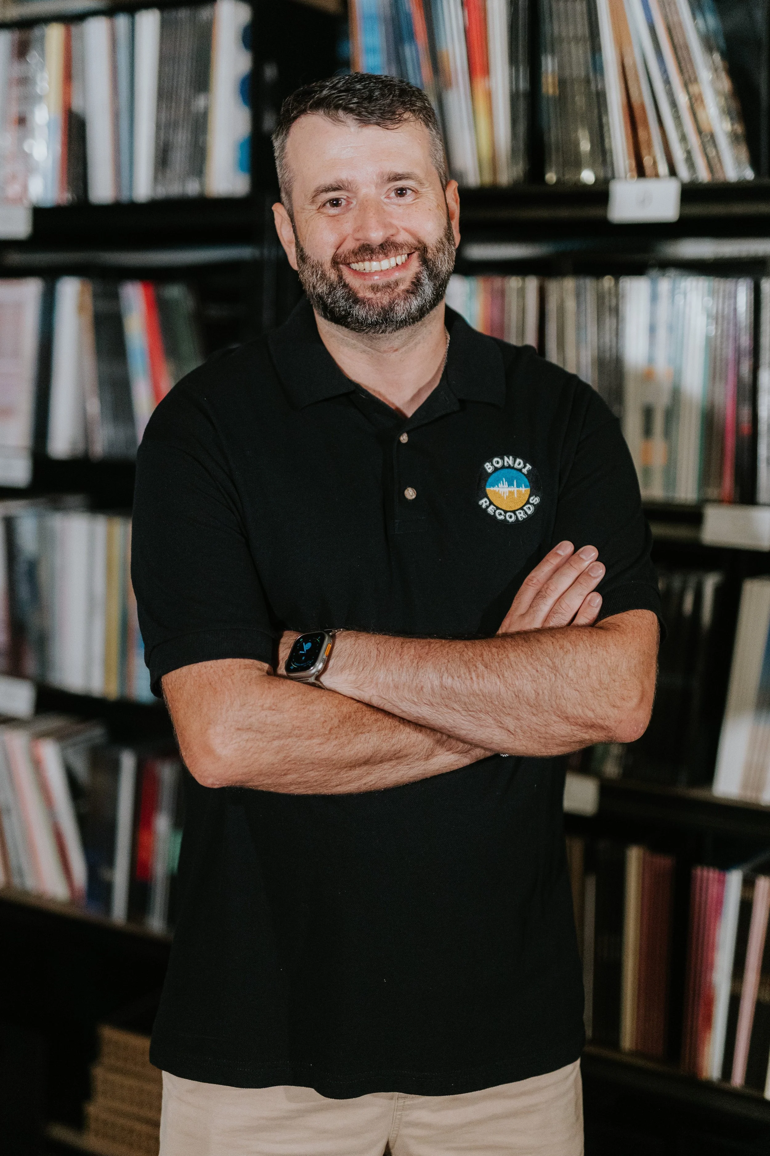 A smiling man with a beard, wearing a black polo shirt with a logo, standing in front of a bookshelf filled with records, arms crossed.