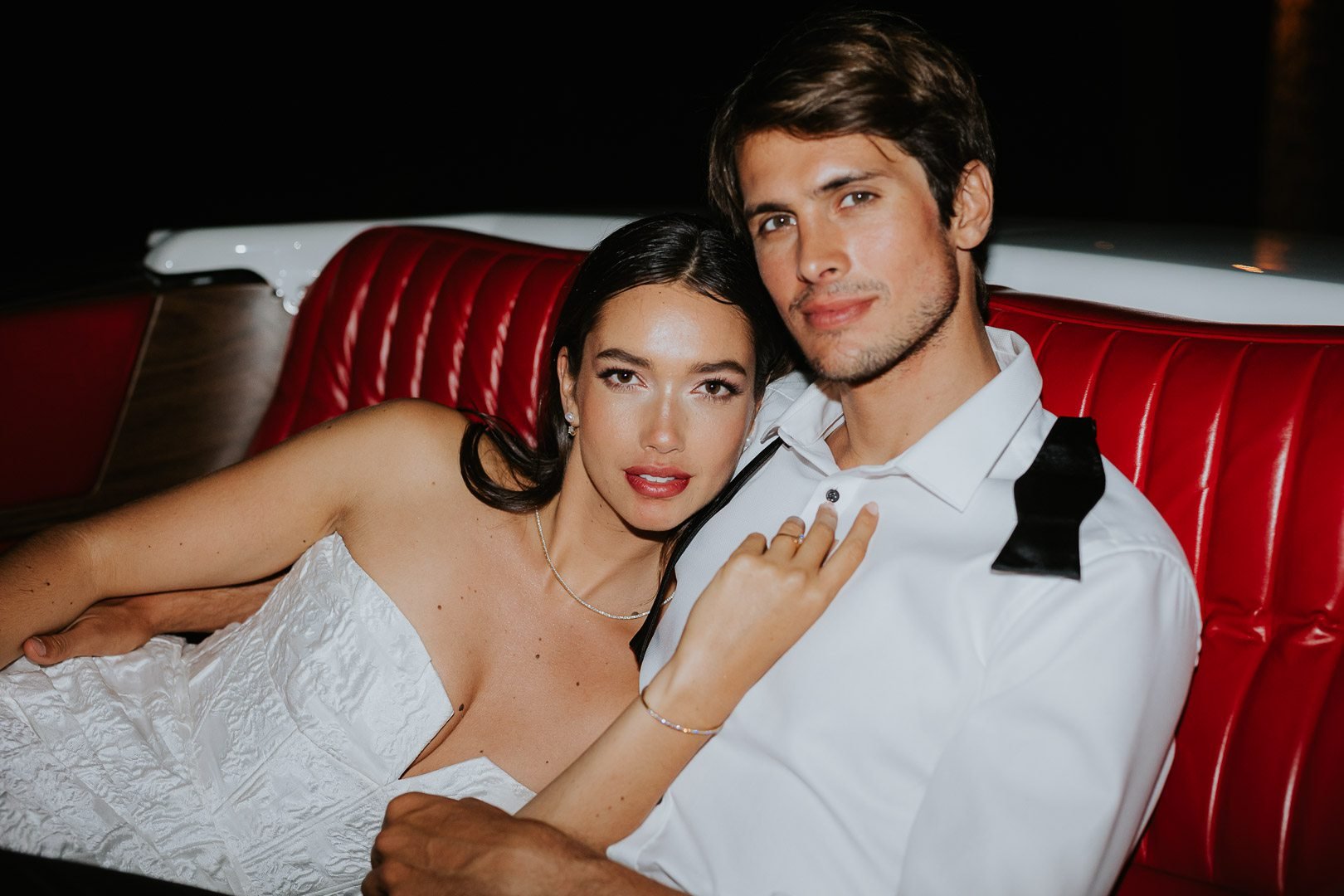 A woman in a white dress and a man in a white shirt with a black bow tie sitting on a red, vintage car seat at night.