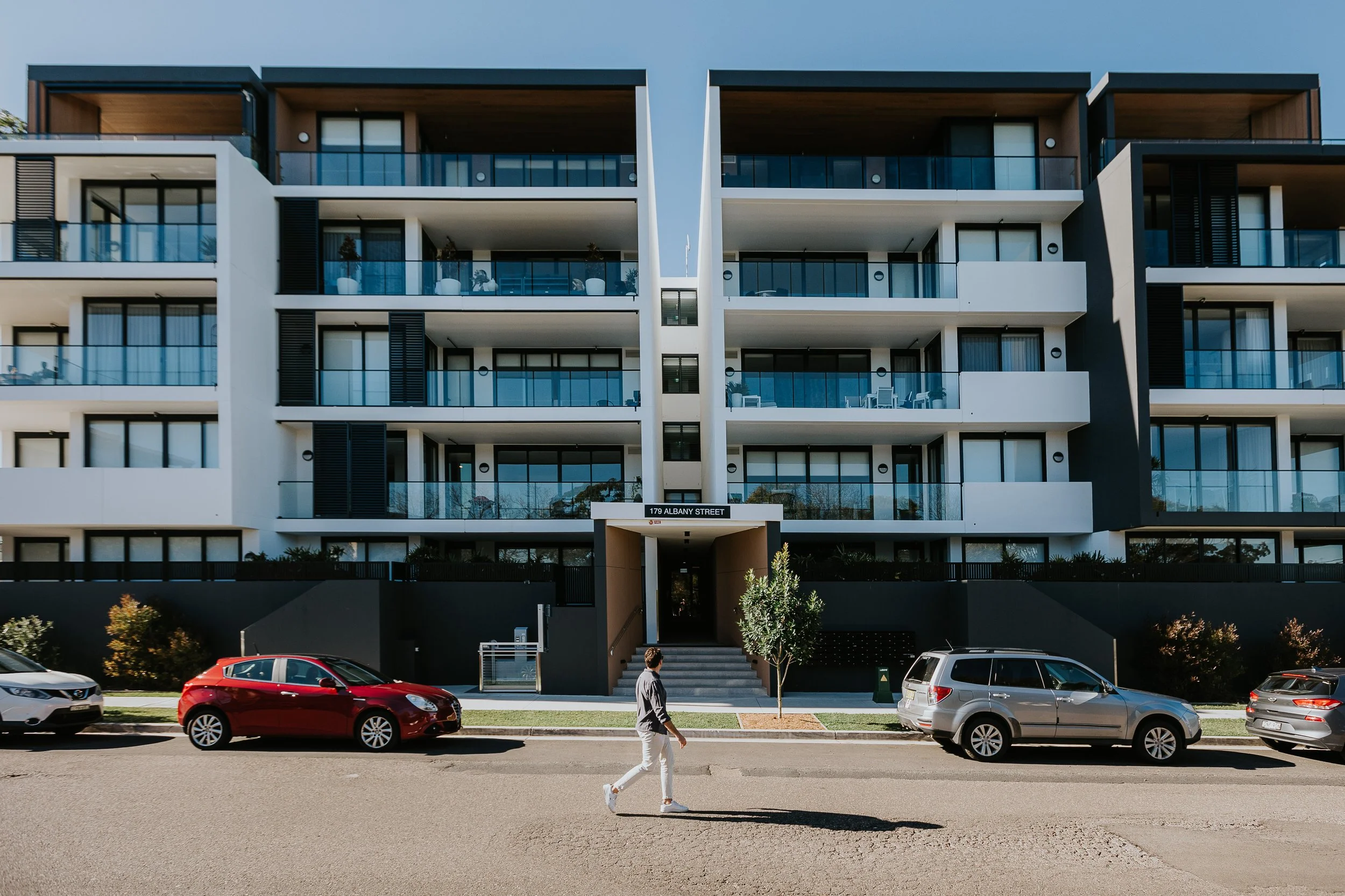 Modern multi-story apartment building with large glass balconies, parked cars in front, and a man walking on the sidewalk.