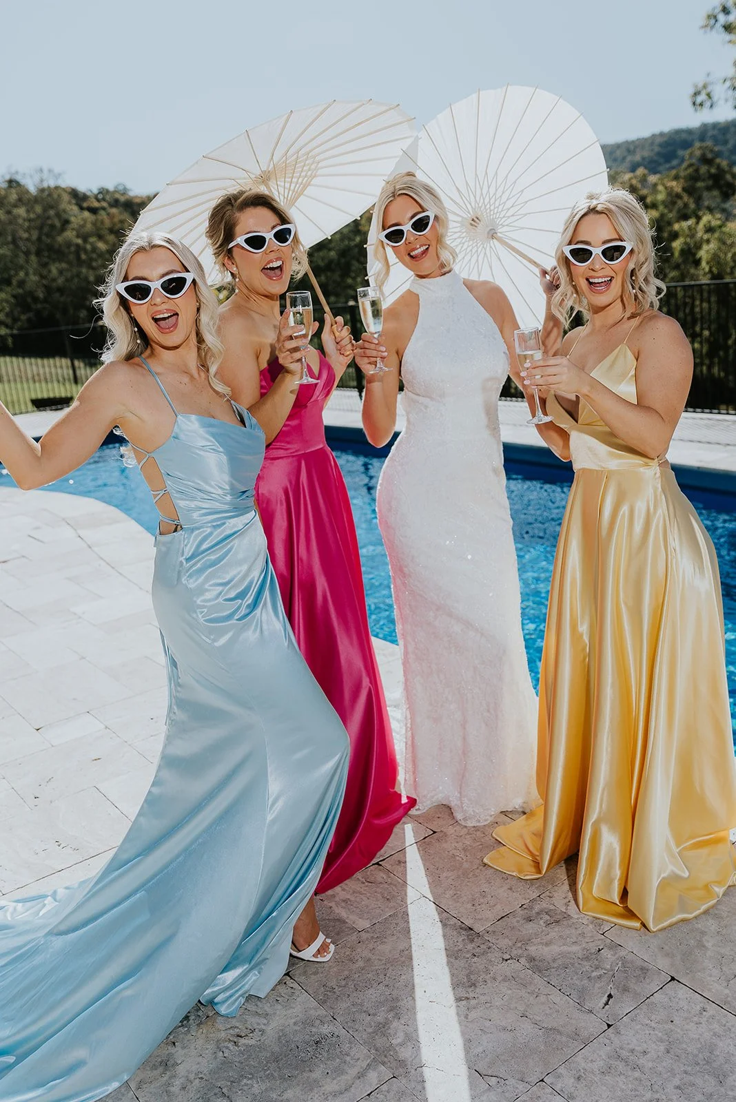 Four women in colorful dresses and sunglasses standing by a pool, holding glasses of champagne and smiling.