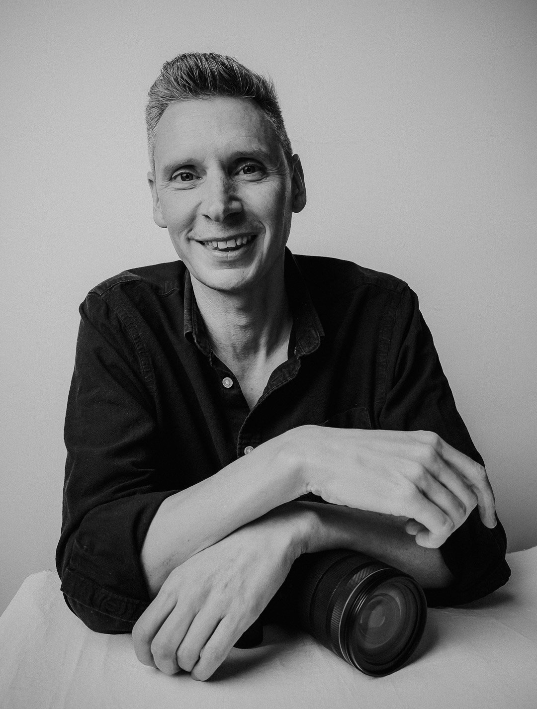 Portrait image of Photographer Christopher Perkins sitting behind a table with arms folded across his camera and smiling. Black & White. Warm and happy.