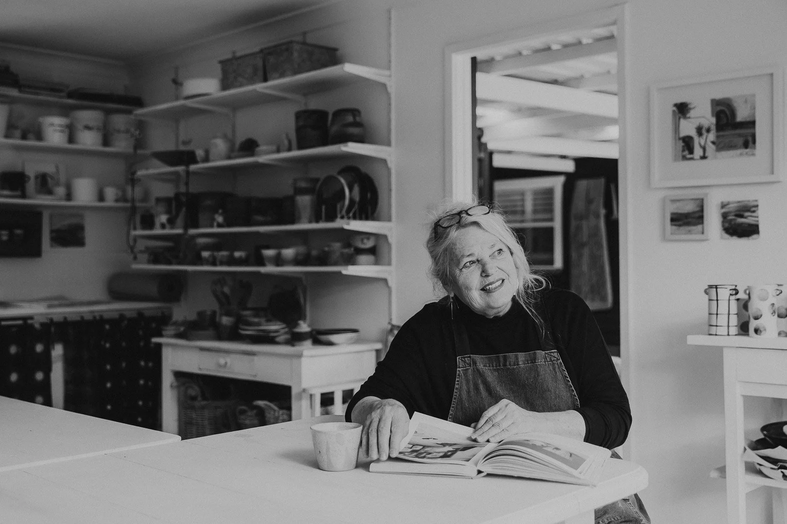 A woman with glasses on her head, smiling, sitting at a kitchen table reading a book, with a coffee mug in front of her. The kitchen has open shelves with dishes and decorative items.