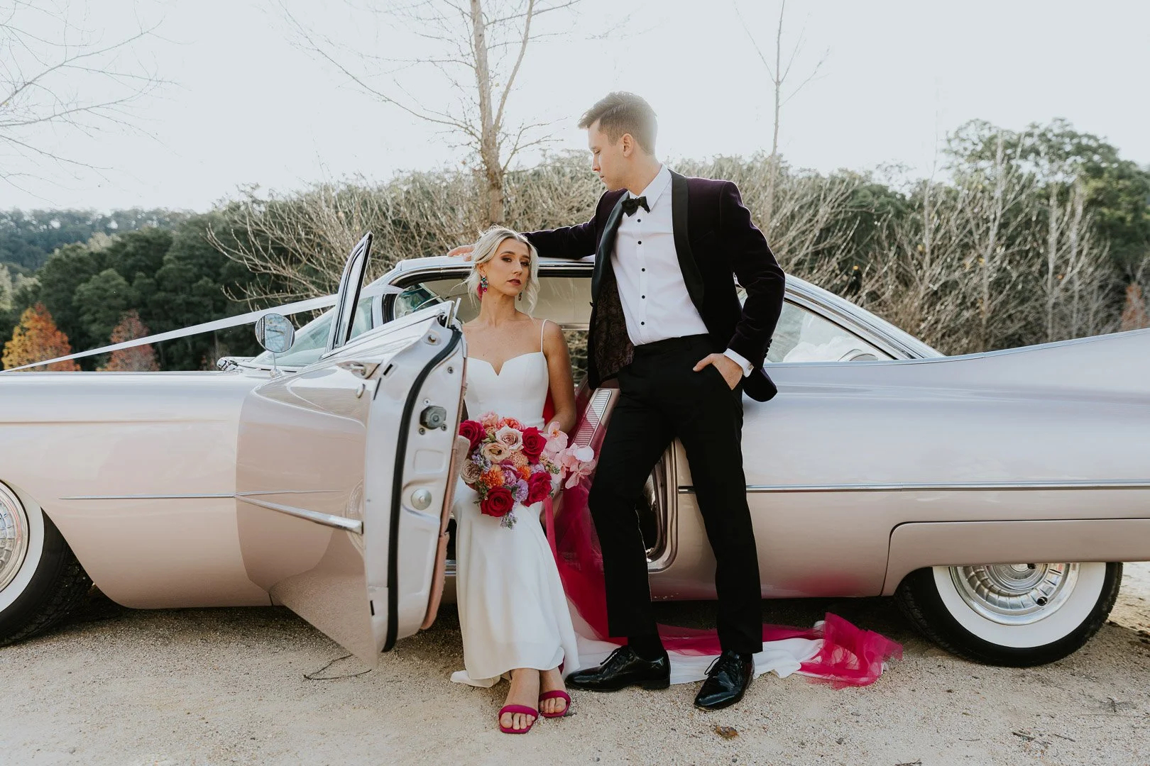 A bride and groom in formal attire posing with a vintage car outdoors. The bride holds a bouquet of pink, red, and white flowers, and the groom rests his hand on the car door while looking at the bride.