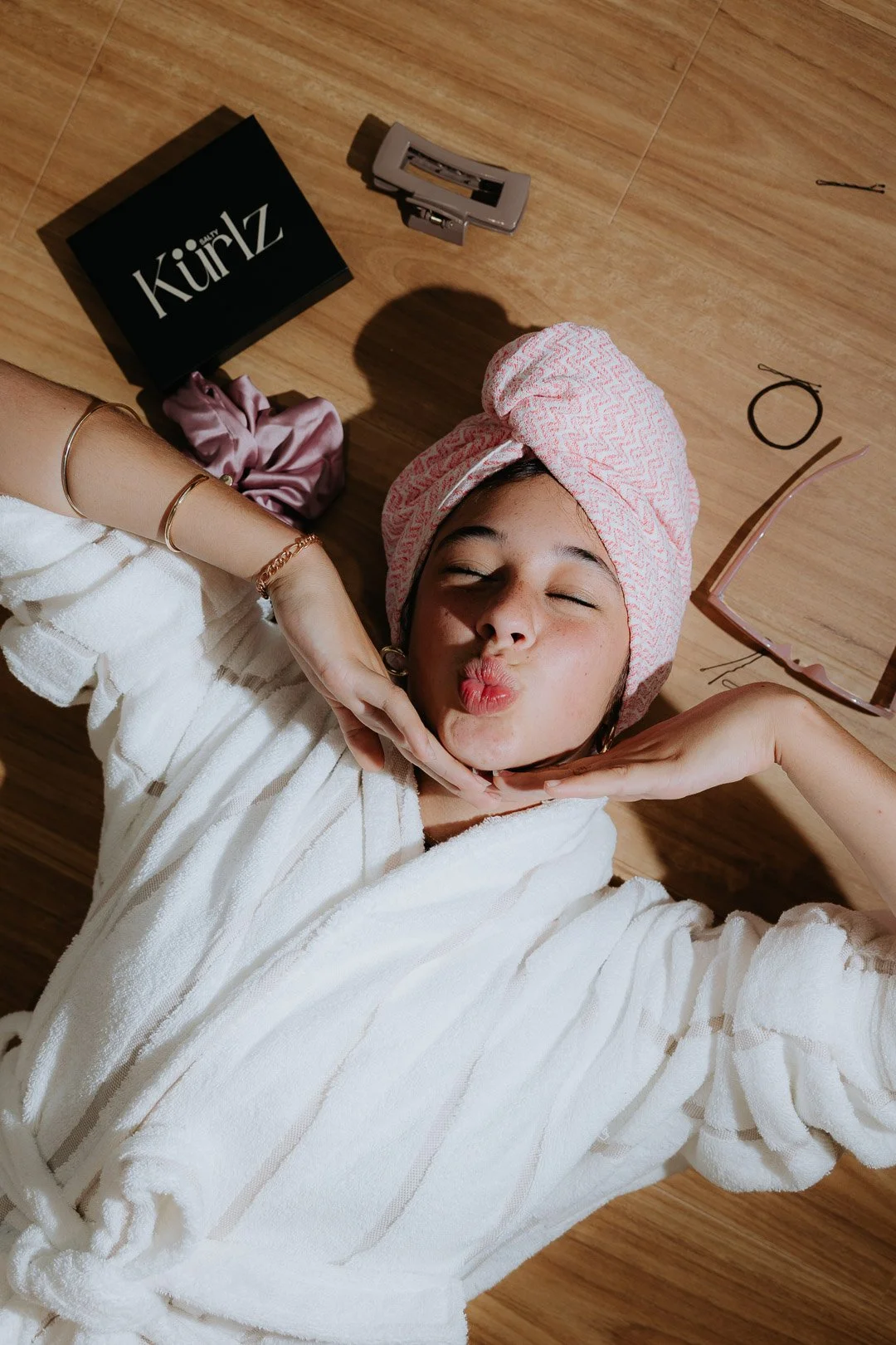 A woman lying on a wooden floor, wearing a pink towel turban and a white bathrobe, making a kissy face while holding her face with her hands. There are sunglasses, a box labeled 'Kimüz,' a hair clip, a pink scrunchie, a small hairbrush, and a black h
