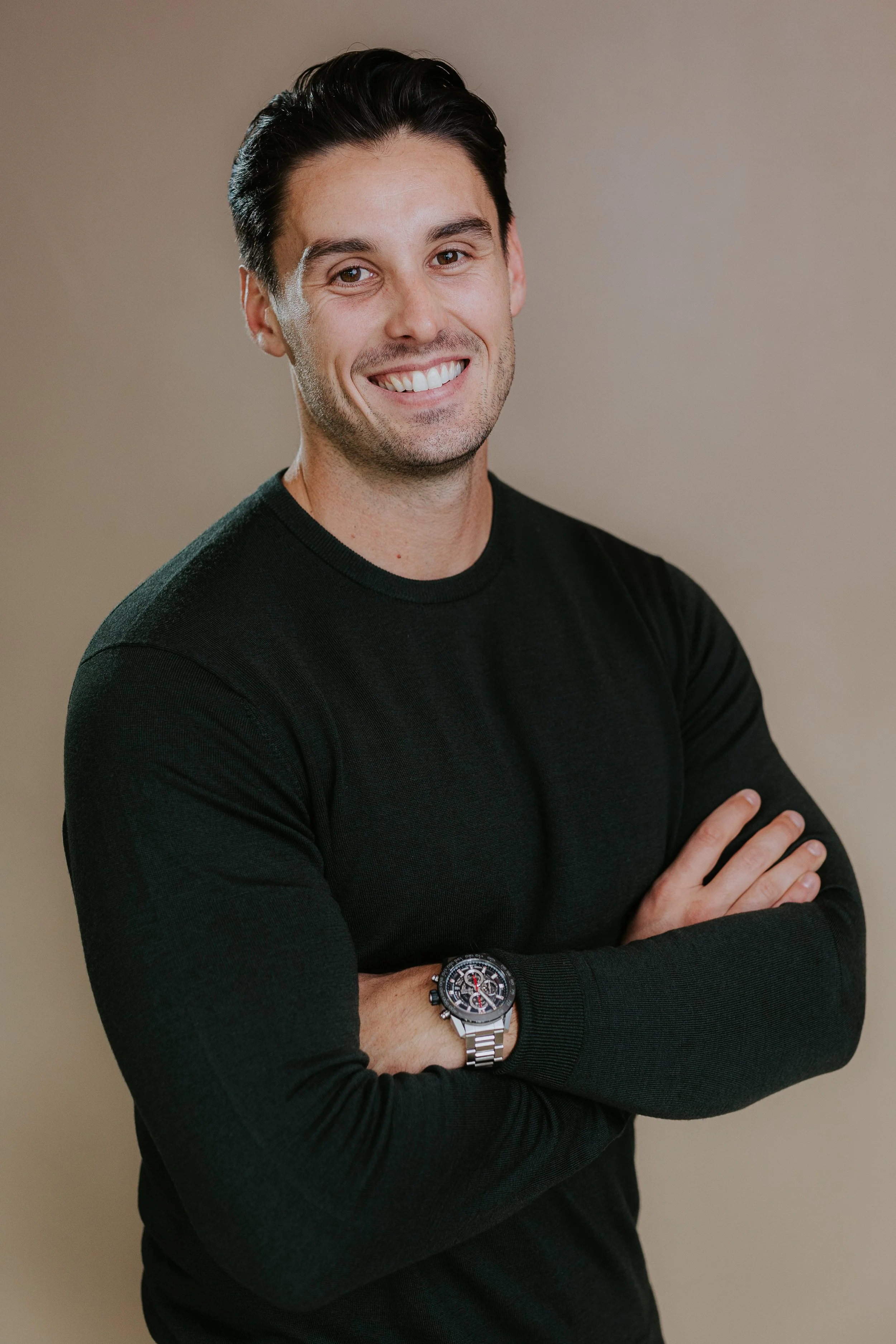 A smiling man with dark hair wearing a black long-sleeve shirt and a large wristwatch, standing against a plain beige background.