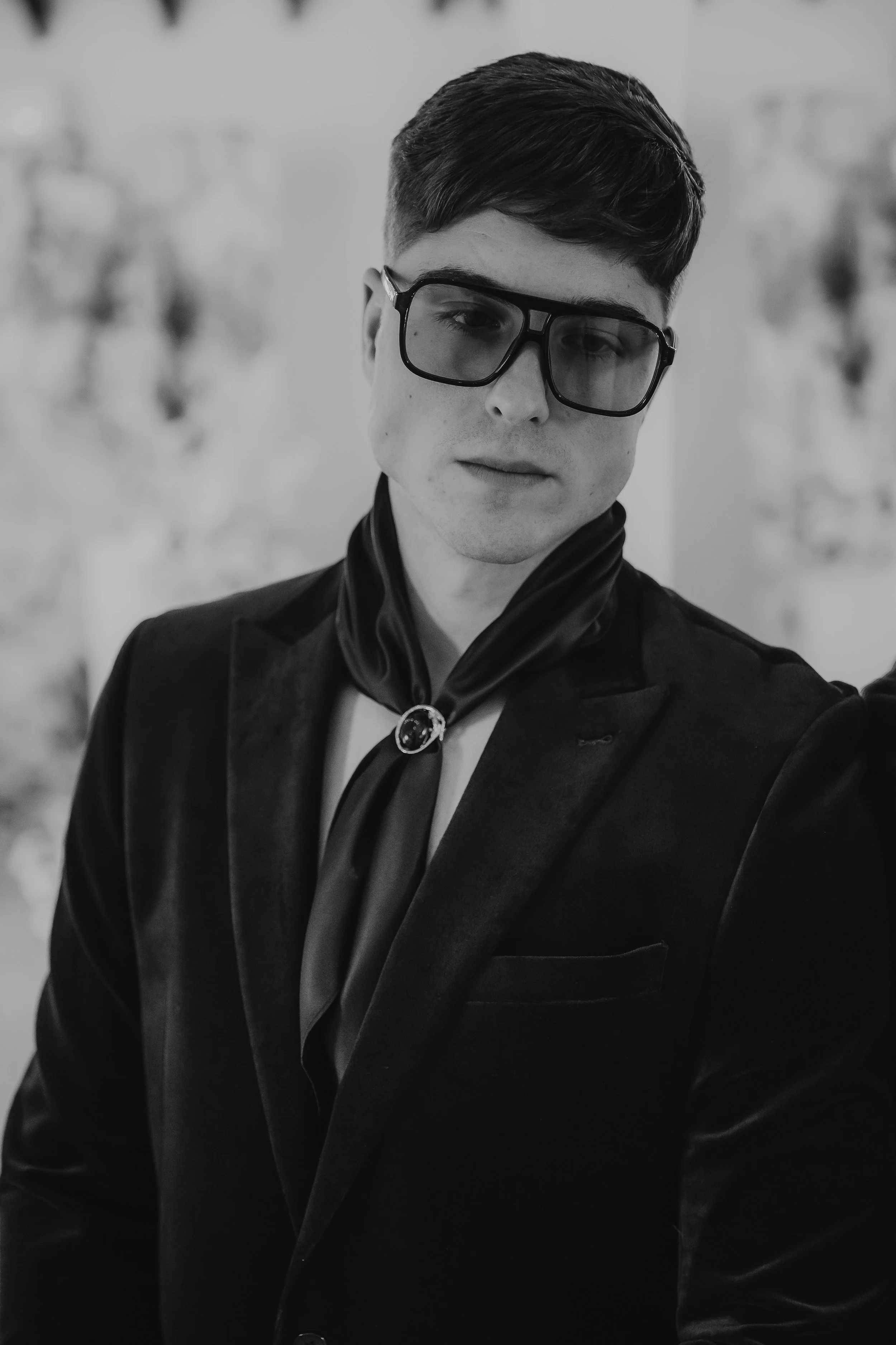 A black-and-white portrait of a young man wearing glasses, a dark blazer, and a silk scarf with a decorative brooch.