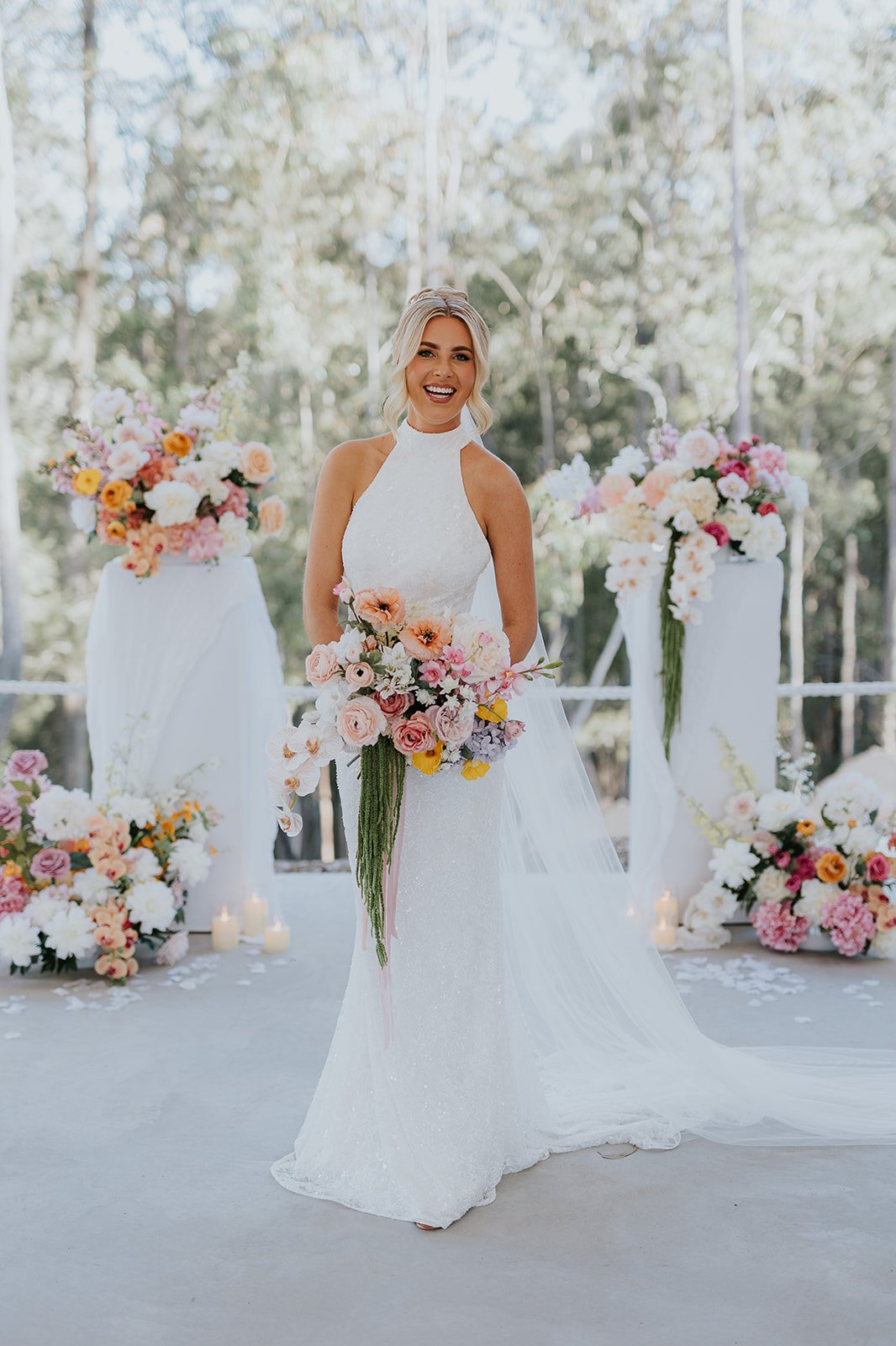 A woman in a white wedding dress holding a bouquet of pink, purple, and yellow flowers, standing outdoors with a backdrop of trees and floral arrangements in the background.