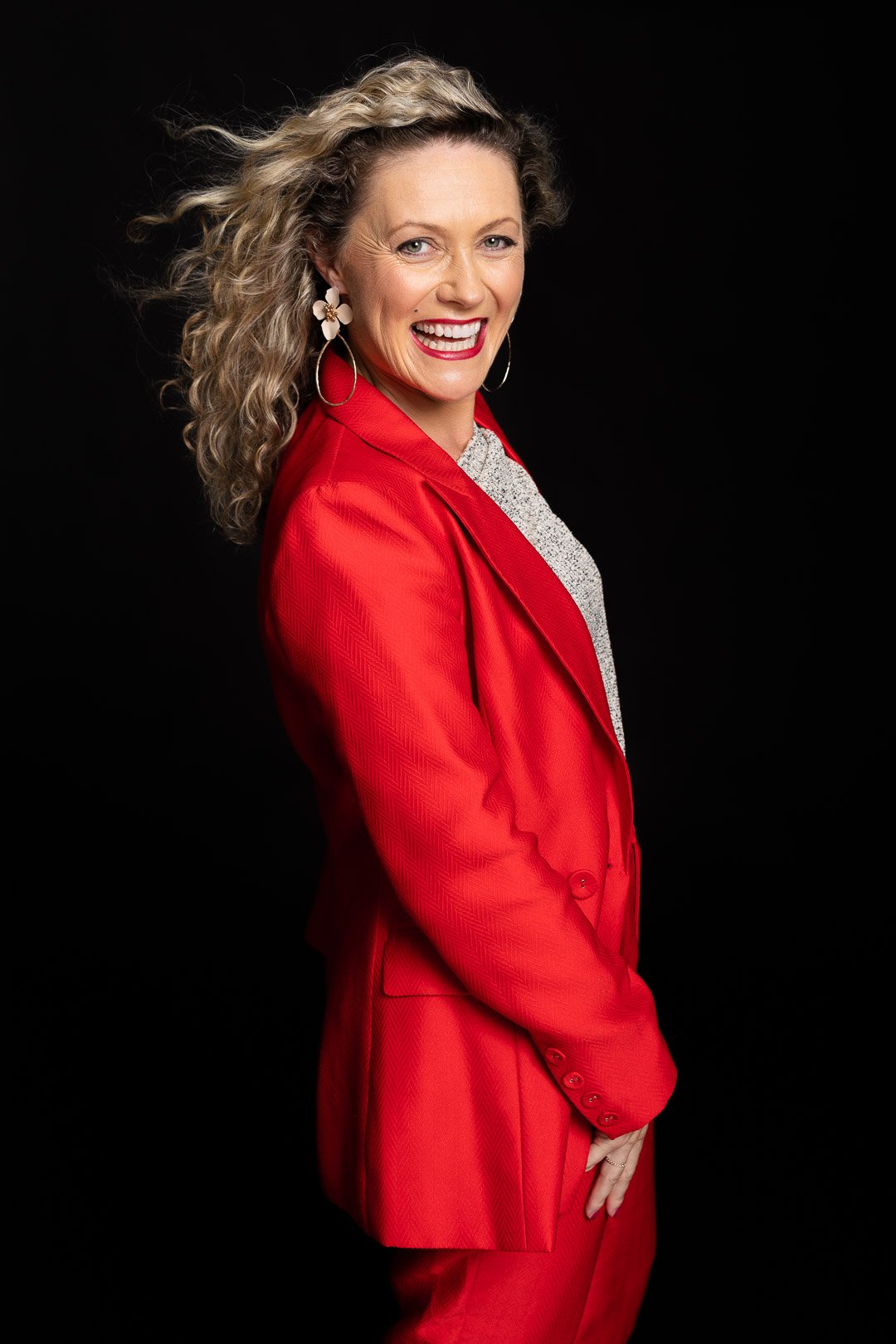 A woman with curly blonde hair smiling, wearing a red blazer, white blouse, and large earrings, posing against a black background.