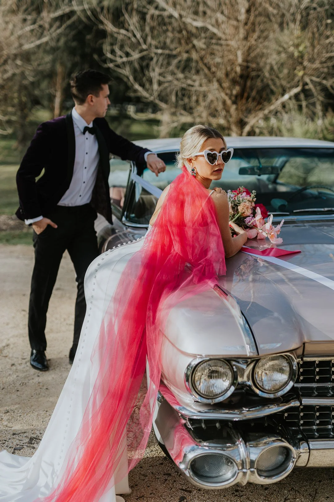 A woman in a wedding dress with a red veil and heart-shaped sunglasses leaning on a vintage silver car decorated with flowers and white ribbon. A man in a tuxedo stands beside her.