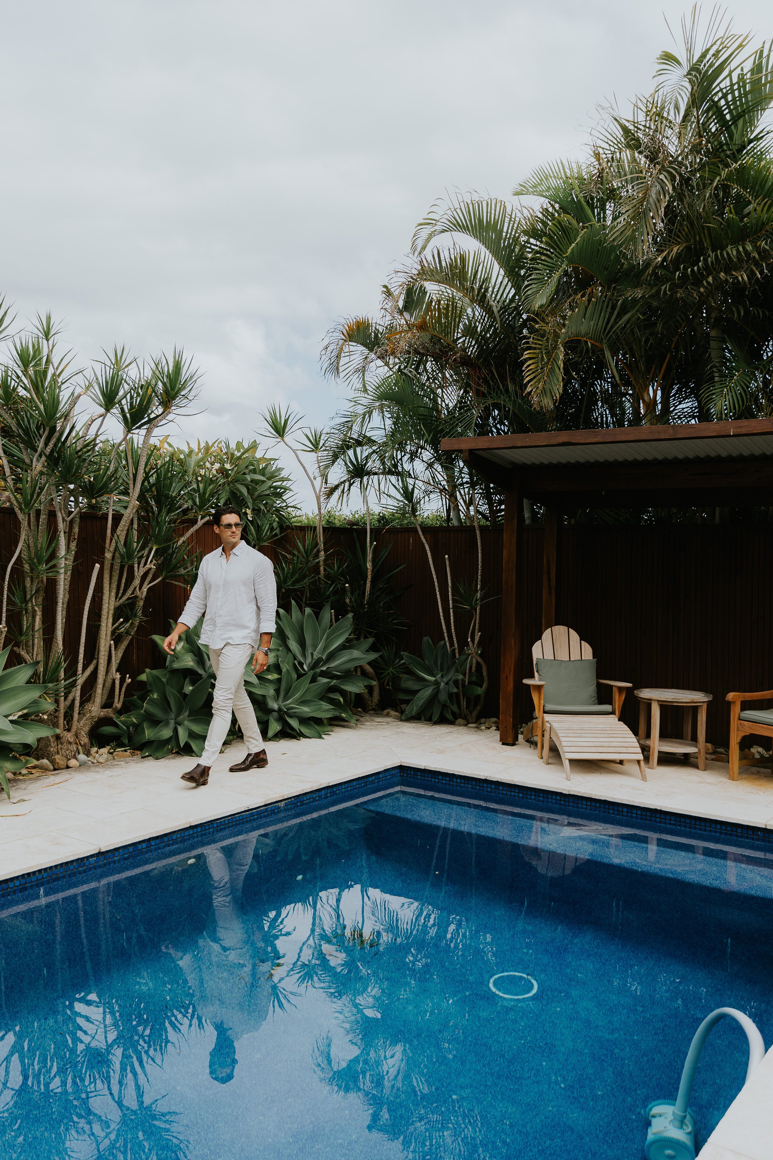 A man dressed in white pants and a shirt walking beside a swimming pool, surrounded by tropical plants and outdoor furniture, under a cloudy sky.