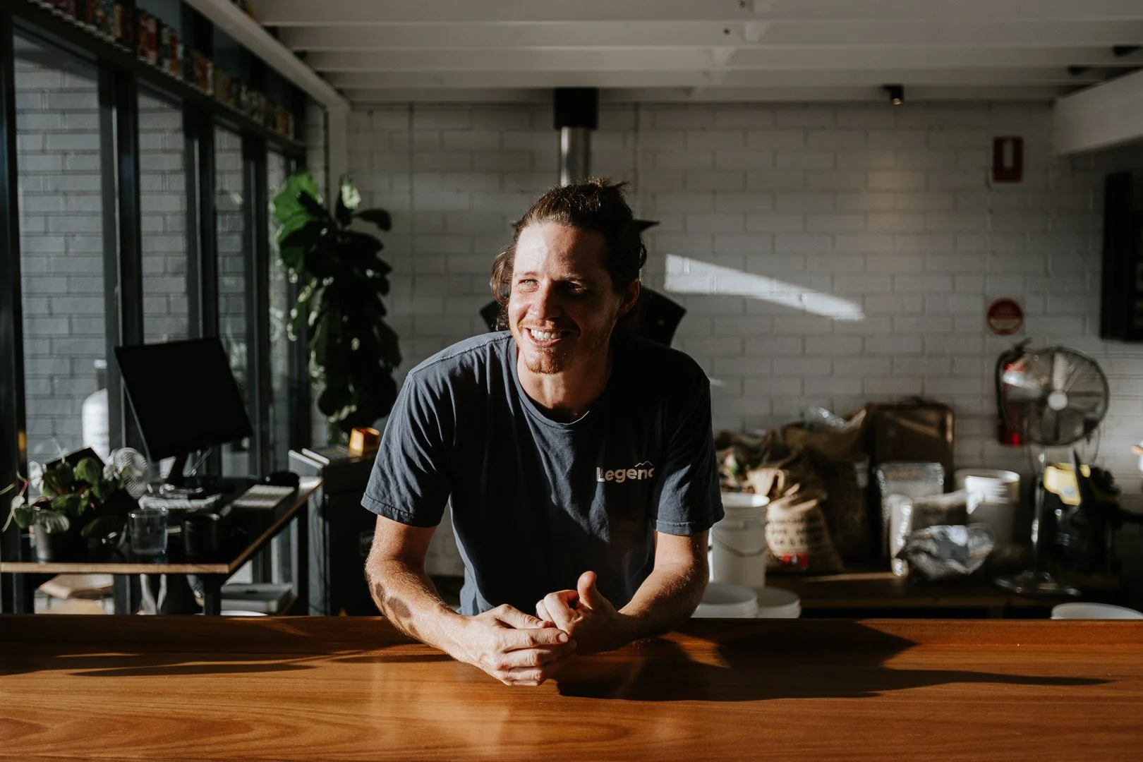 A smiling man with dark hair tied back, wearing a dark t-shirt, leaning on a wooden counter in a bright, modern room with white brick walls, large windows, and various kitchen items in the background.
