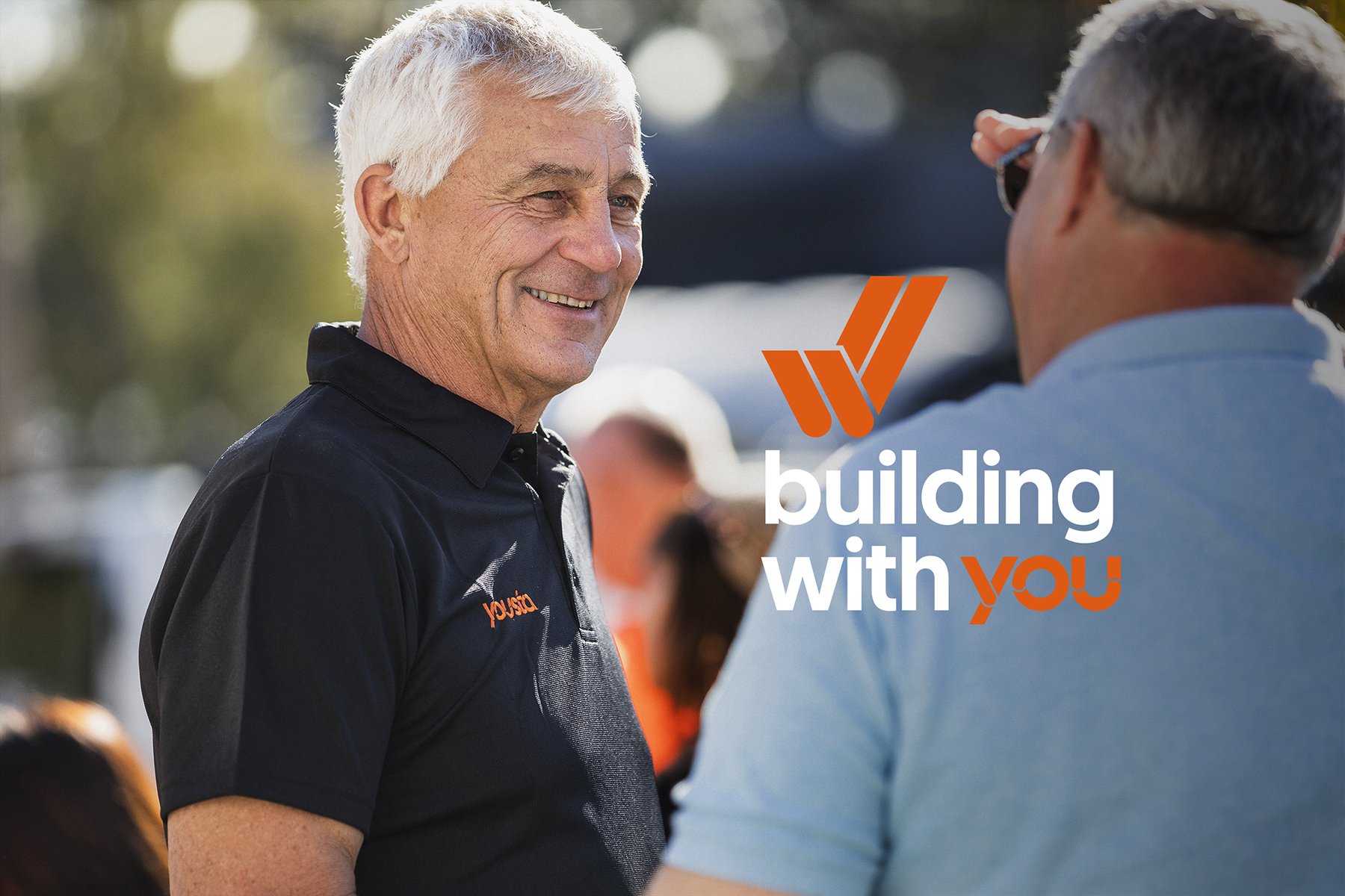 Two men smiling and talking outdoors, with the text 'building with YOU' overlayed. One man wears a black polo with a logo, and the other wears a light blue shirt.