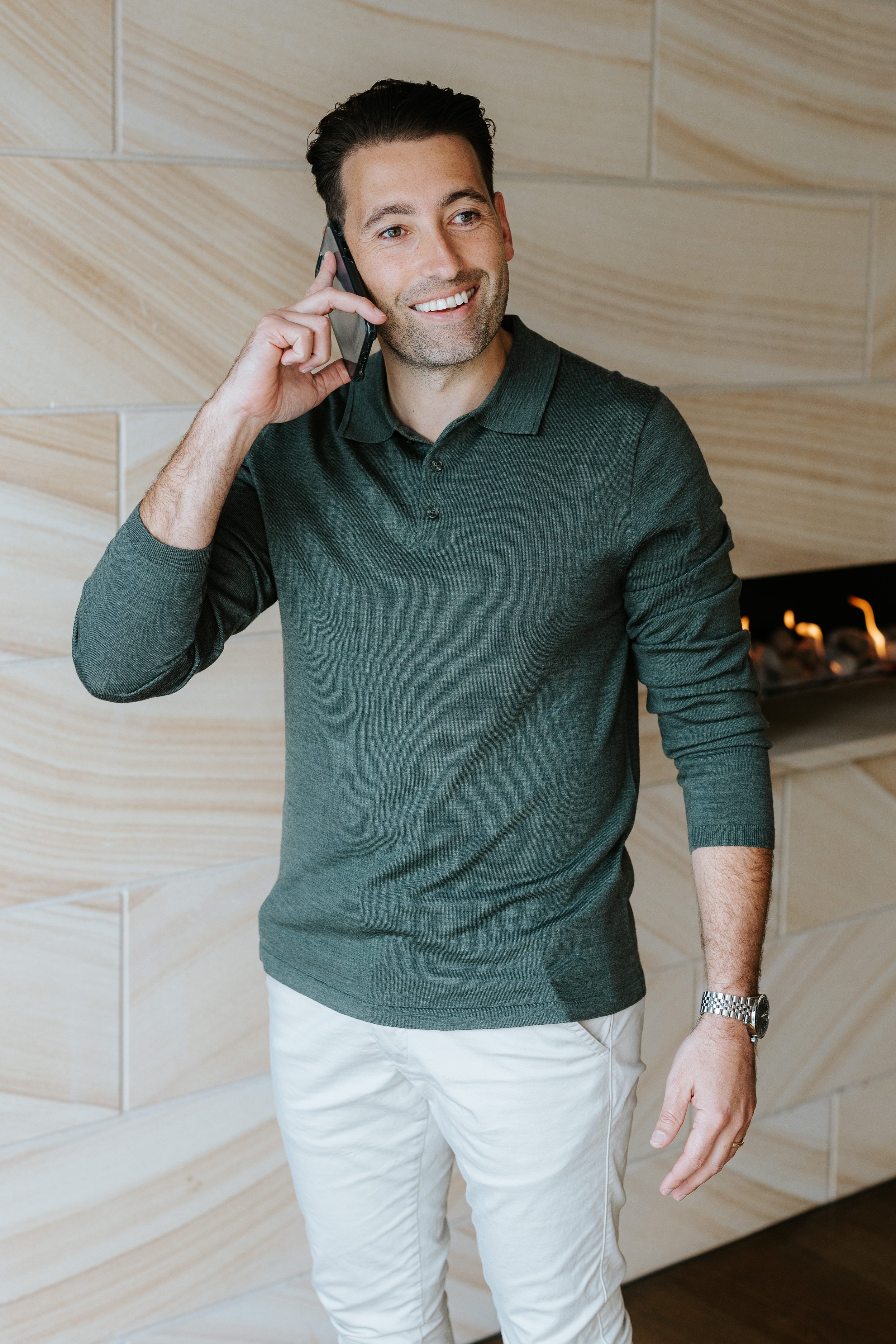 A man smiling and talking on a smartphone indoors with a modern wooden wall and fireplace in the background.