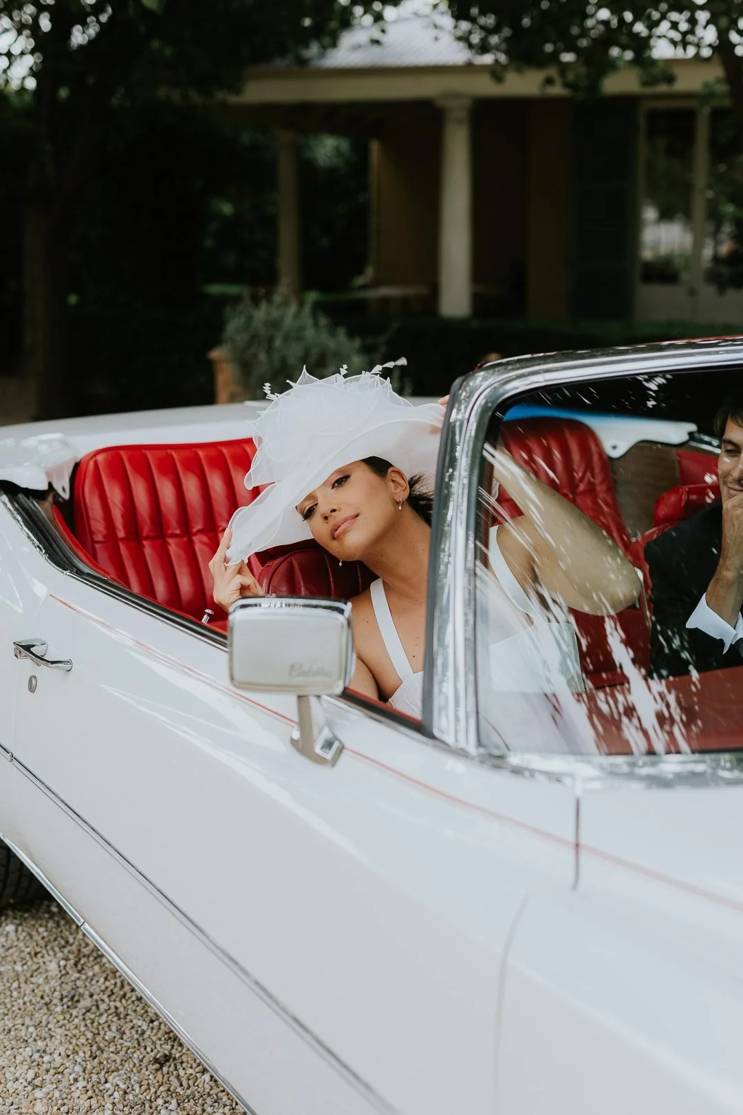 Woman wearing a large white hat, sitting in a vintage white convertible car with red interior, smiling at camera.