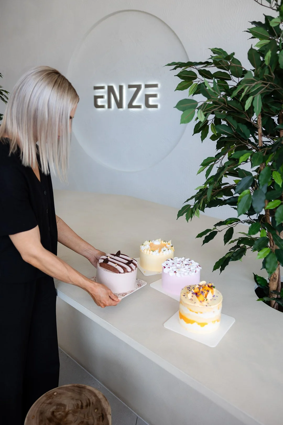 A woman with blonde hair arranging four decorated cakes on a white table in a modern, minimalistic space with a large green plant and a sign that reads 'ENZE' on the wall.
