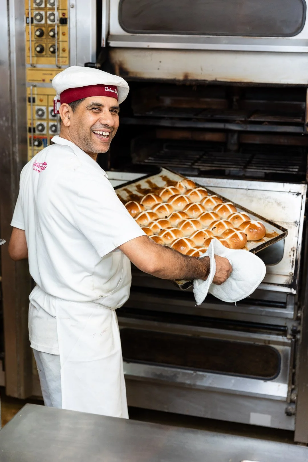 A man wearing a white chef's uniform and a red and white chef's hat with a name embroidered on it is taking freshly baked bread rolls out of an industrial oven. He is smiling and holding a baking tray with bread rolls.