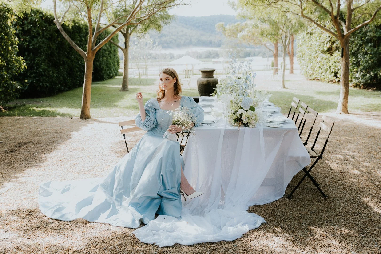 A woman in a blue gown sitting at an outdoor table with white tablecloth, floral arrangements, and chairs, surrounded by trees in a park-like setting.