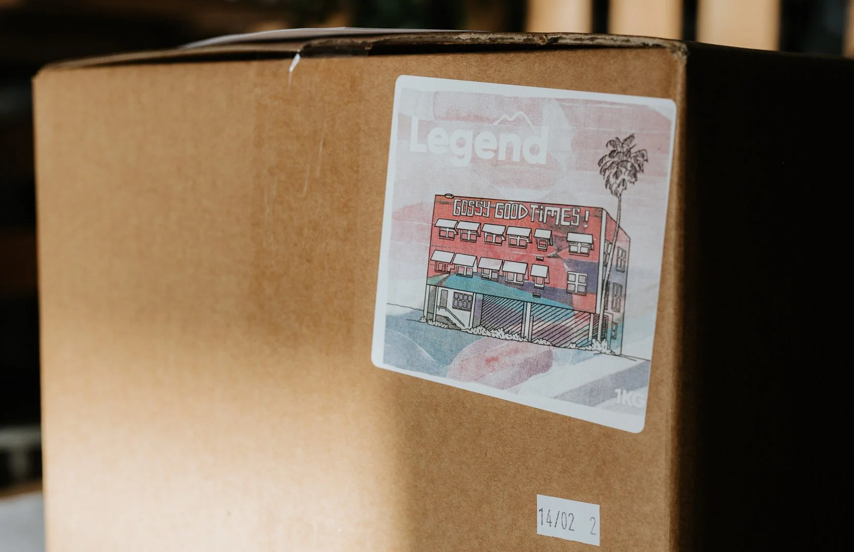A close-up of a cardboard box with a sticker featuring an illustrated building and the text "Gossy Good Times!" above it. The sticker has a white border and some text at the top, partially visible, that reads "Legend."