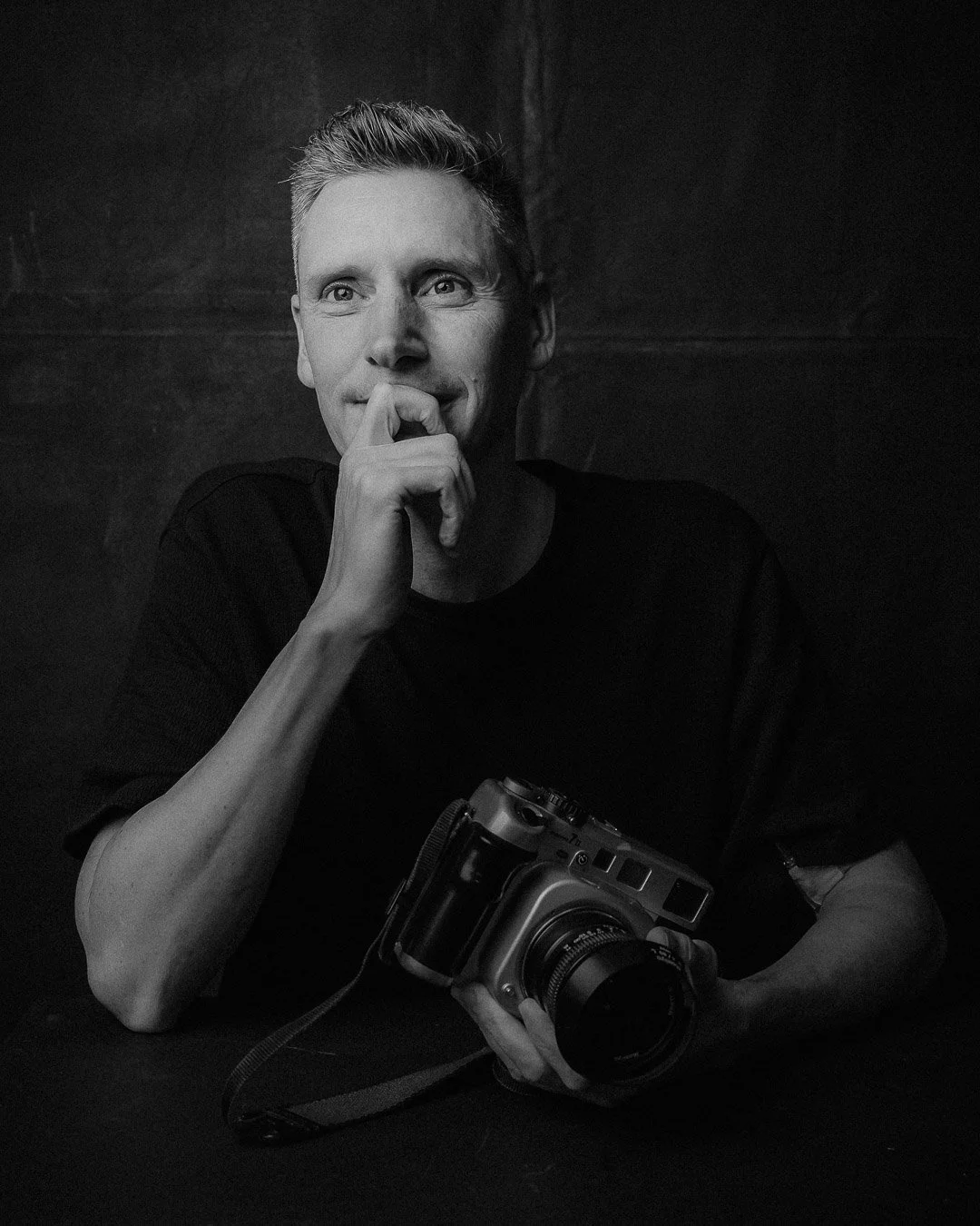 Black & White Studio Portrait Photo of Christopher Perkins, Australian Lifestyle and Travel Photographer holding a camera, smiling and looking off camera
