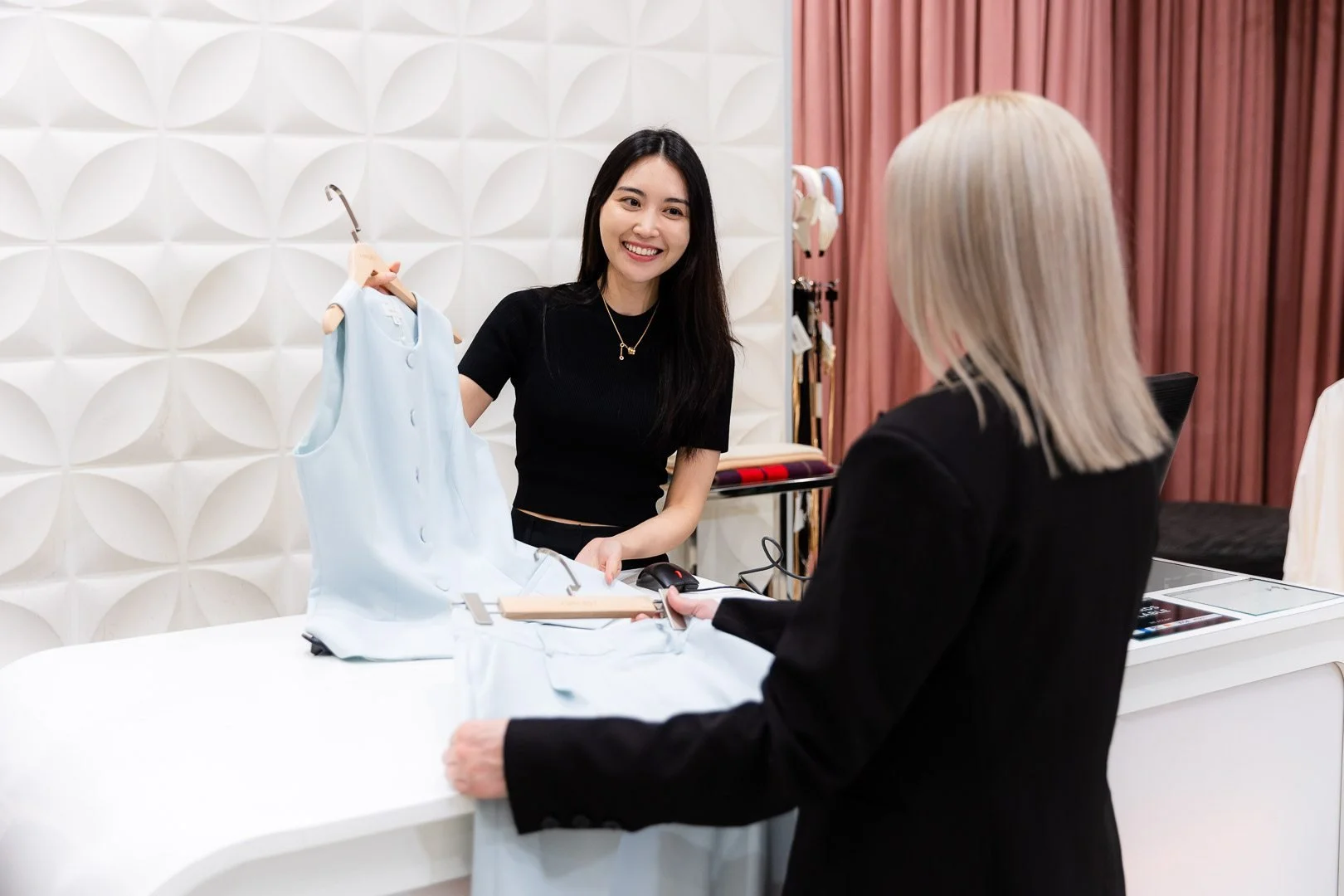 A woman at a retail counter showing a light blue dress to a customer with blonde hair in a black blazer, inside a boutique with textured white wall and pink curtains.