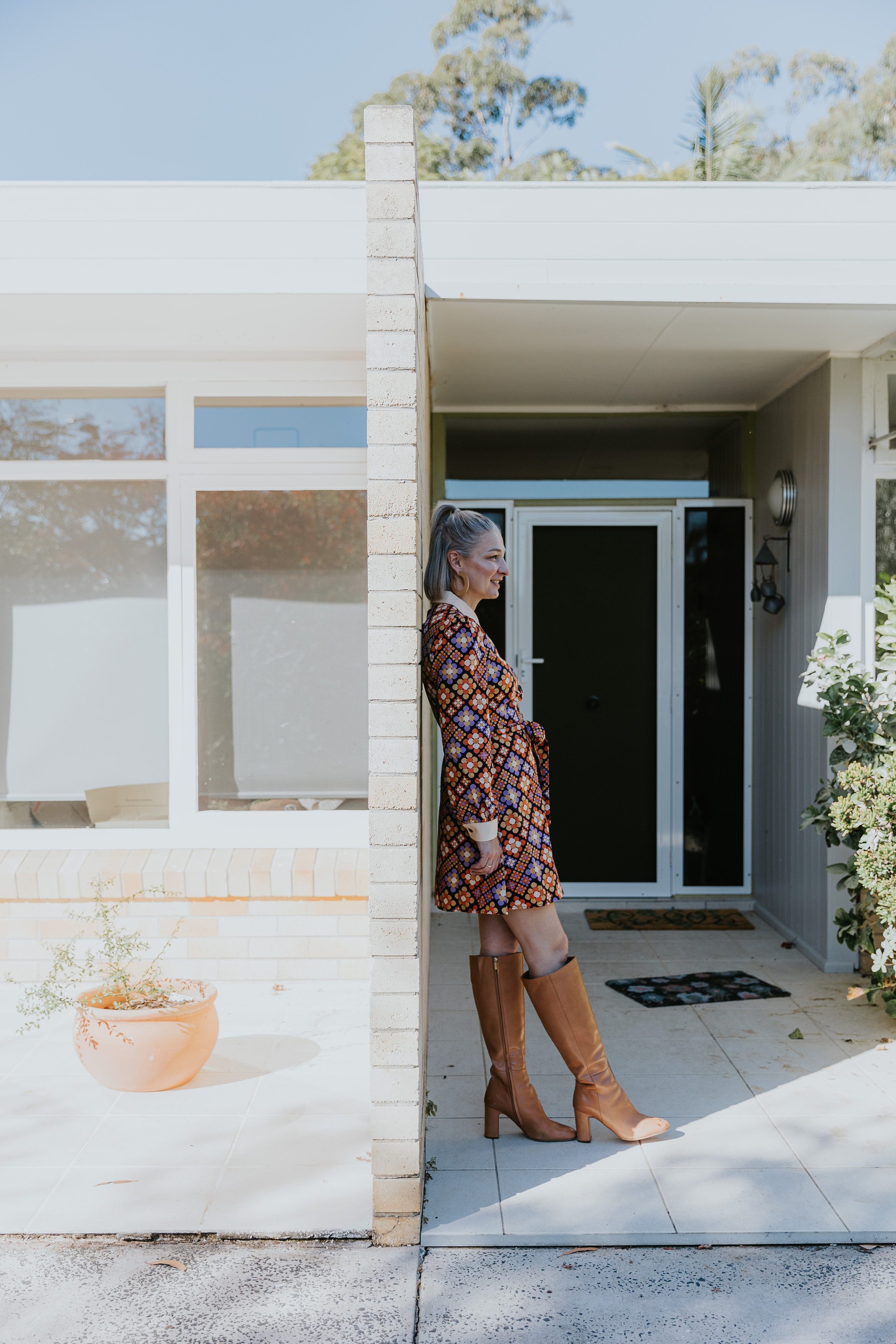 Woman standing on front porch, leaning against exterior wall, wearing a patterned dress and tall brown boots.