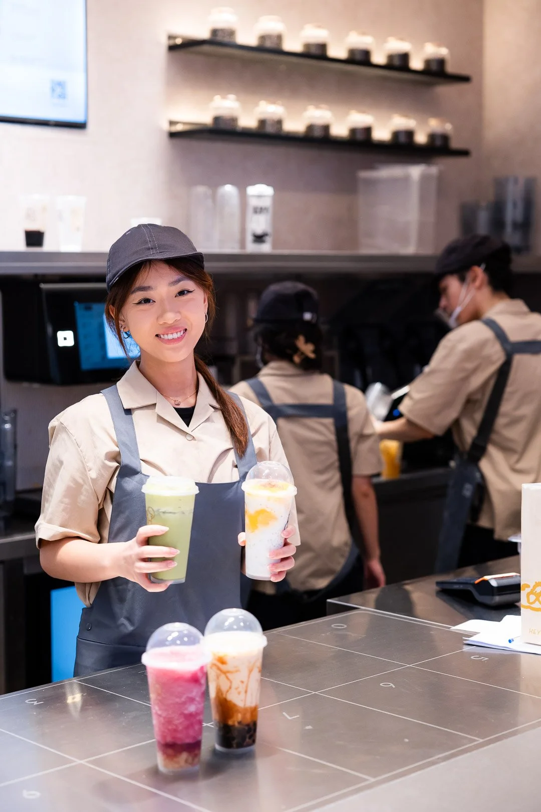 A smiling barista holding two drinks, standing behind a counter with three colorful beverages, with other staff working in the background at a coffee shop.