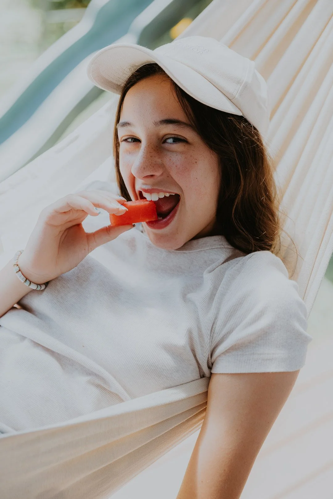 Young woman in a white cap and shirt sitting on a hammock, eating a slice of watermelon and smiling.